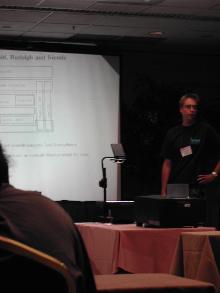 A presenter stands beside a projector, giving a talk on JVM with a slide displayed on the screen.
