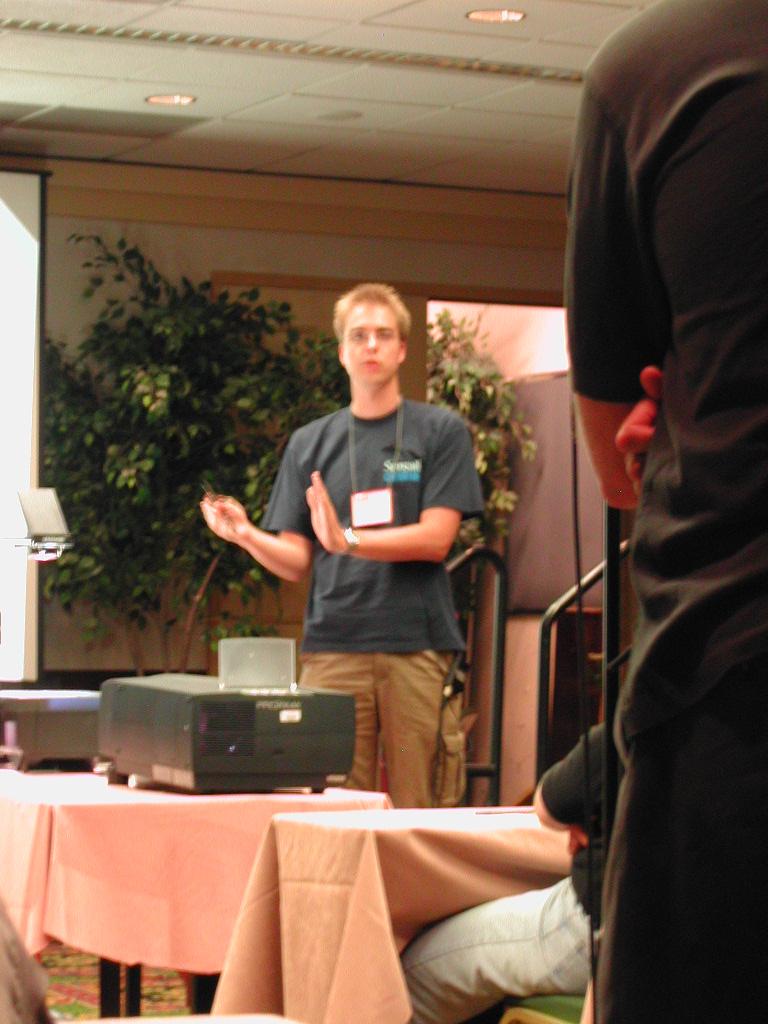 A presenter in a dark shirt gestures while speaking at a conference, with an audience member in the foreground.