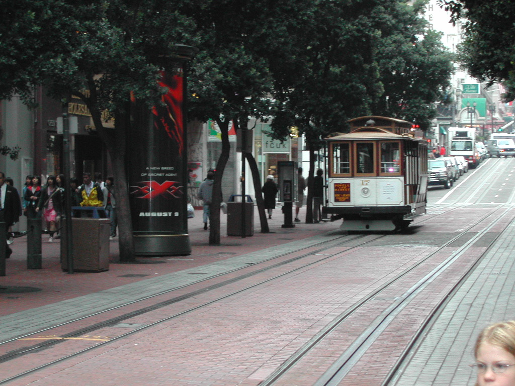 A San Francisco cable car moves along tracks in a busy street, surrounded by pedestrians and city buildings.