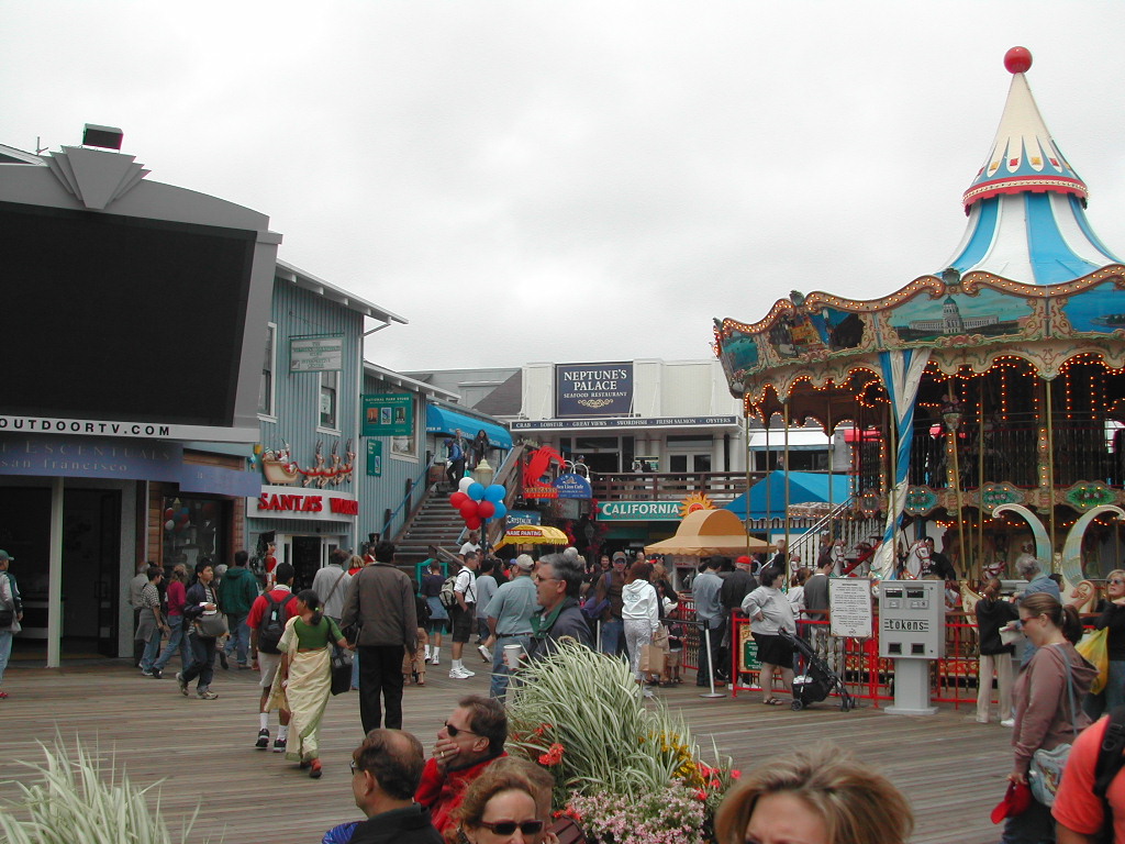 A busy pier with people walking, a colorful carousel, and various shops and restaurants in the background.