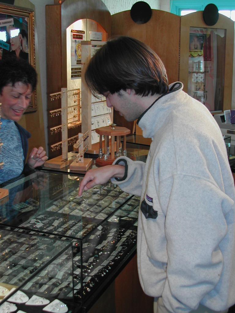 A man in a light-colored jacket points at jewelry inside a glass display case while talking to a salesperson.