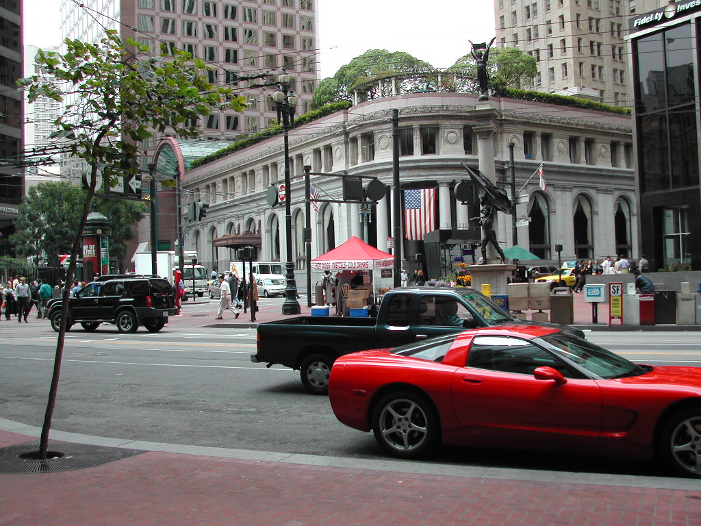 A busy San Francisco street with cars, pedestrians, and a historic building with flags and a small red tent.