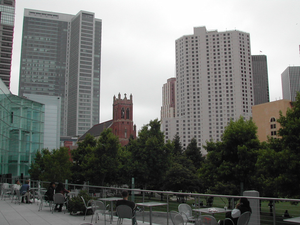 People sit at outdoor tables in a public space with tall buildings and a church in the background.