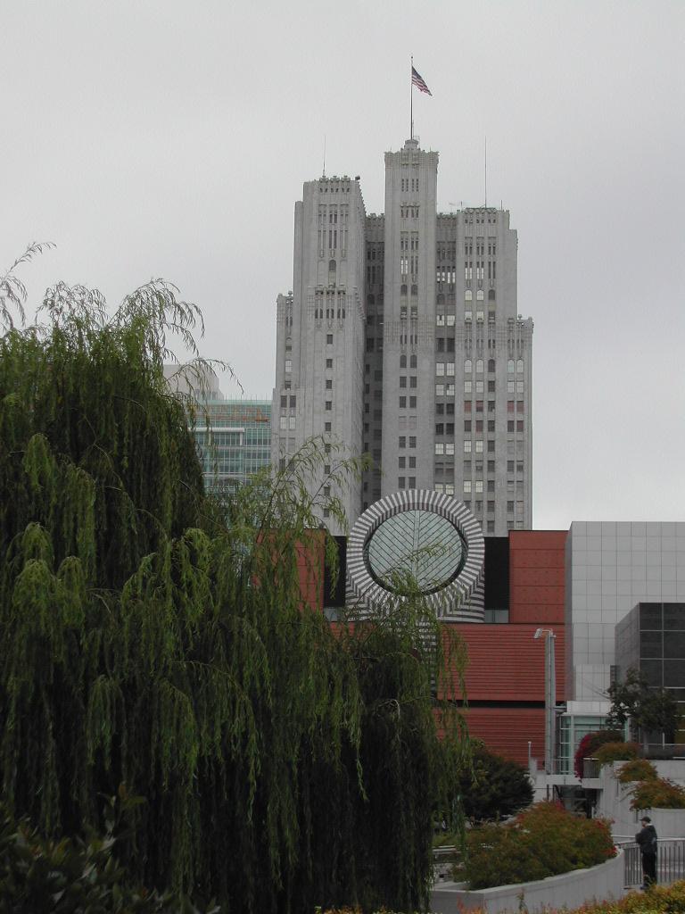 A tall building with an American flag behind the San Francisco Museum of Modern Art, partially obscured by trees.
