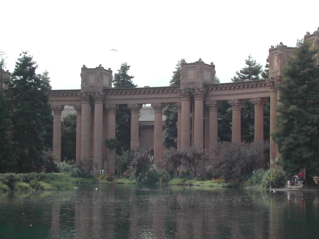 A large classical-style colonnade with tall columns reflected in a pond, surrounded by trees and greenery.