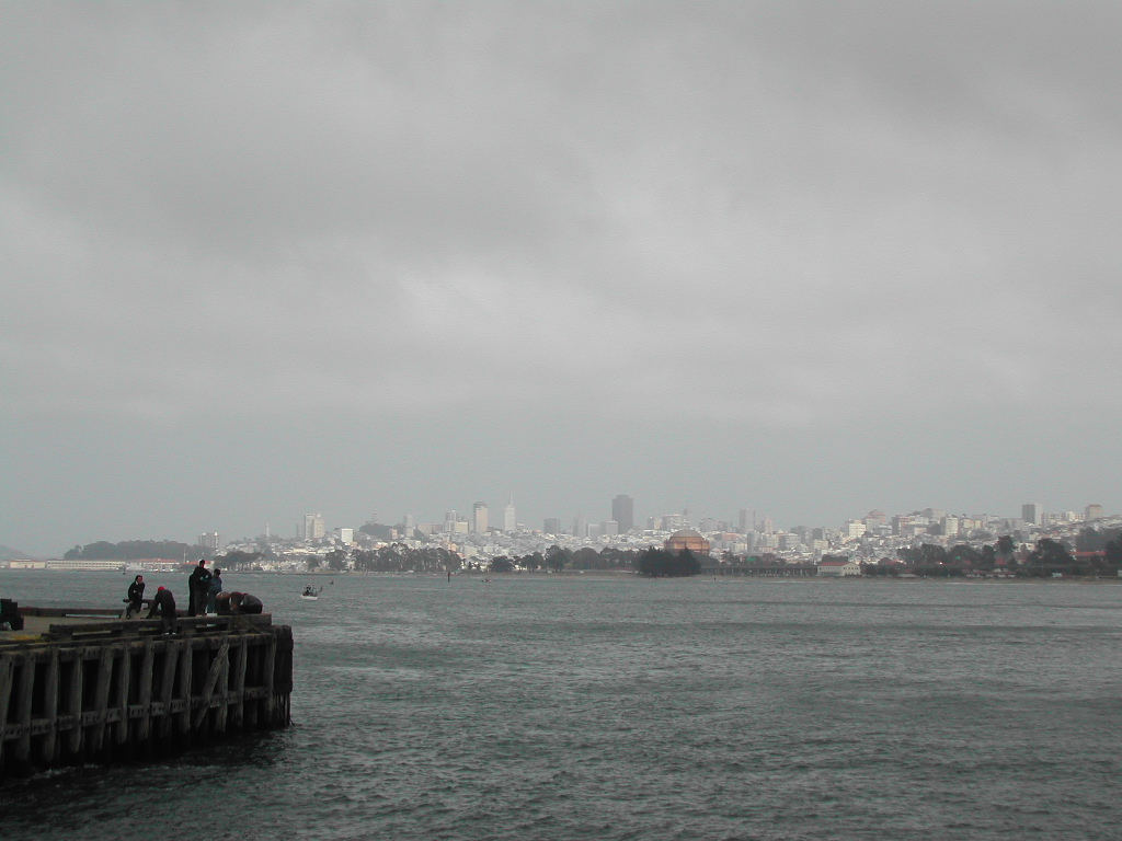 People fishing on a wooden pier with the San Francisco skyline in the background under a cloudy sky.