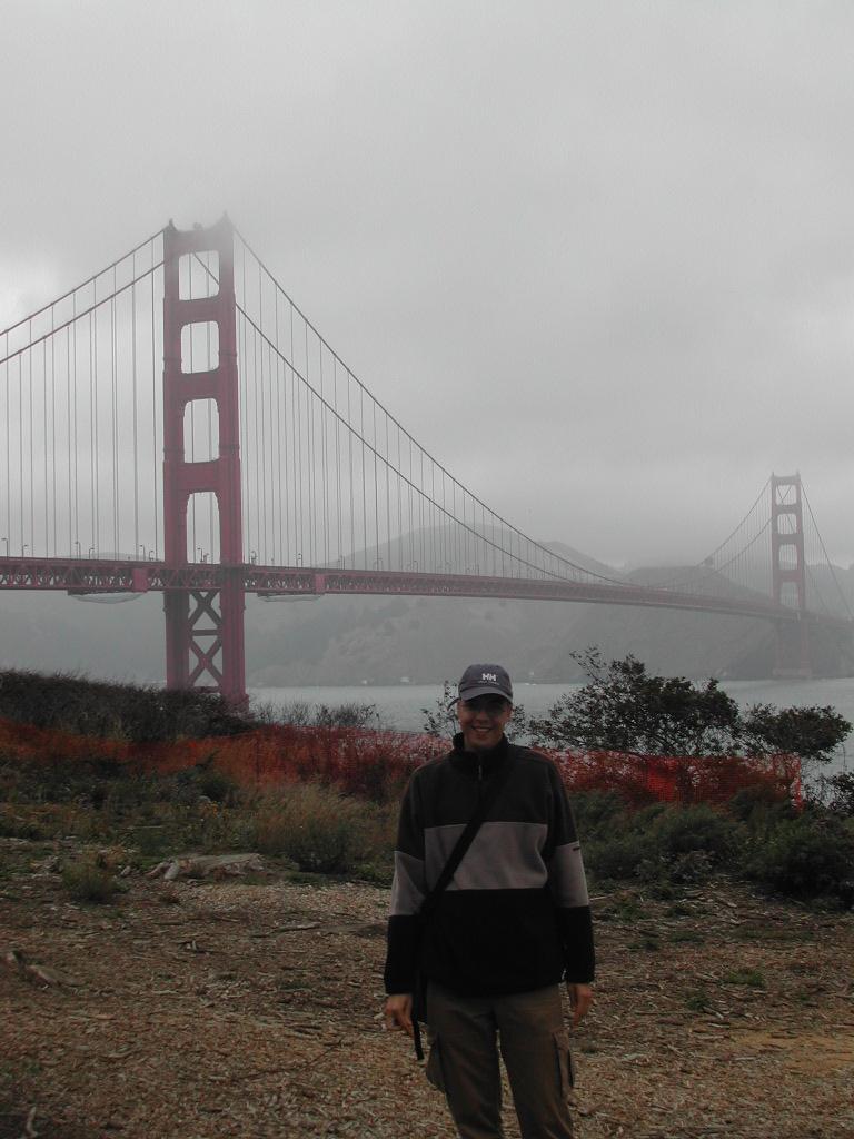 A person wearing a jacket and cap stands in front of the Golden Gate Bridge on a foggy day.
