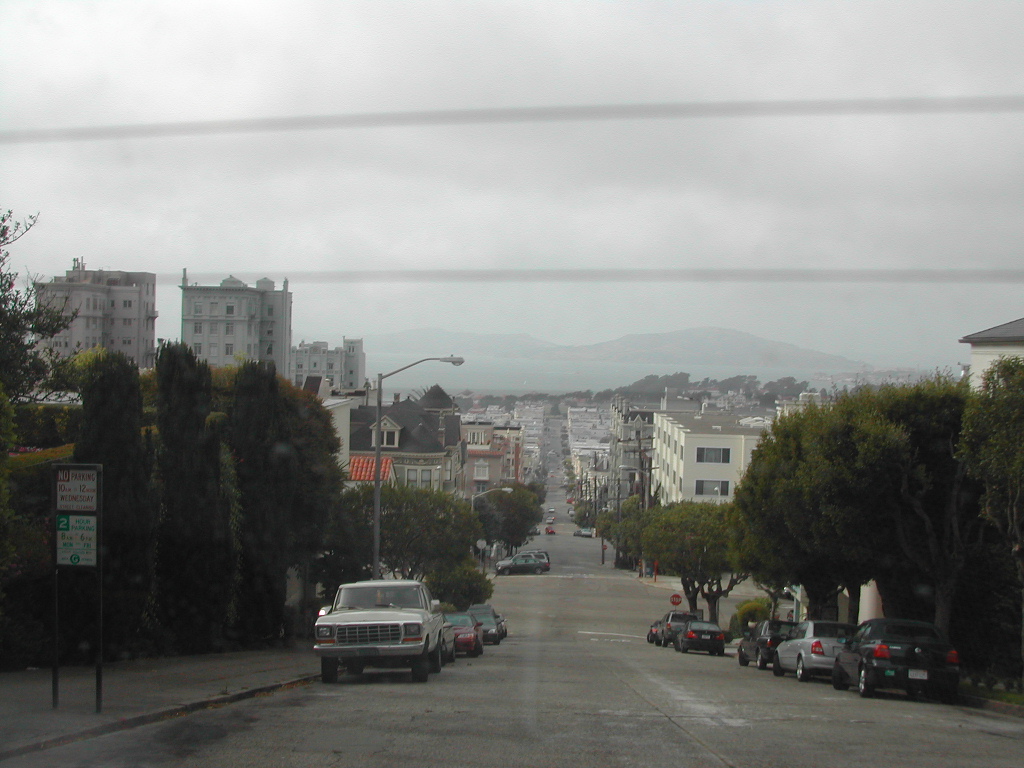 A steep street in San Francisco with parked cars, buildings, and a cloudy sky in the background.
