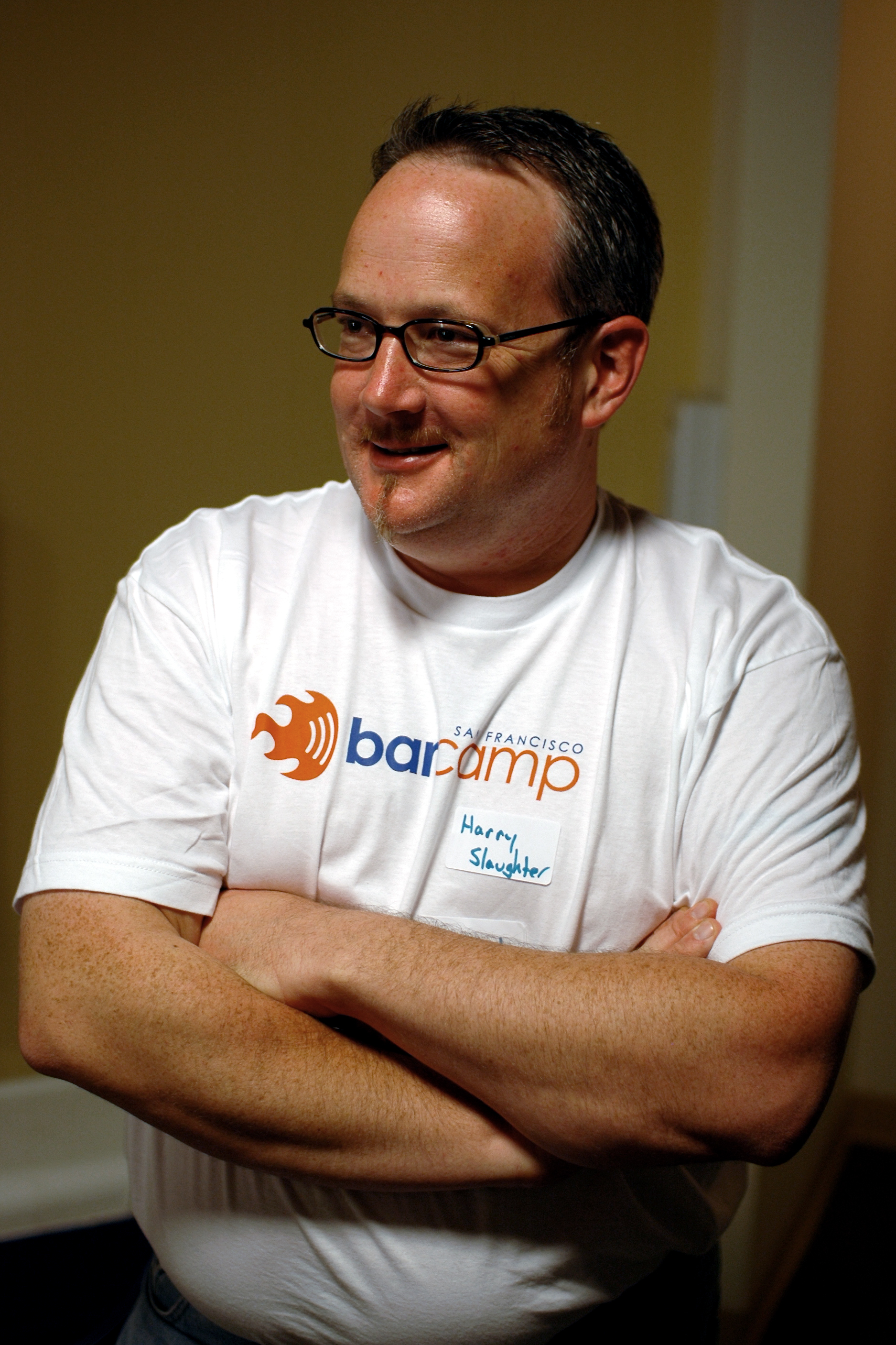 A man wearing a BarCamp San Francisco t-shirt and name tag stands with arms crossed, smiling.