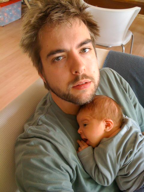 A tired-looking man sits on a couch, holding a newborn baby resting on his chest.