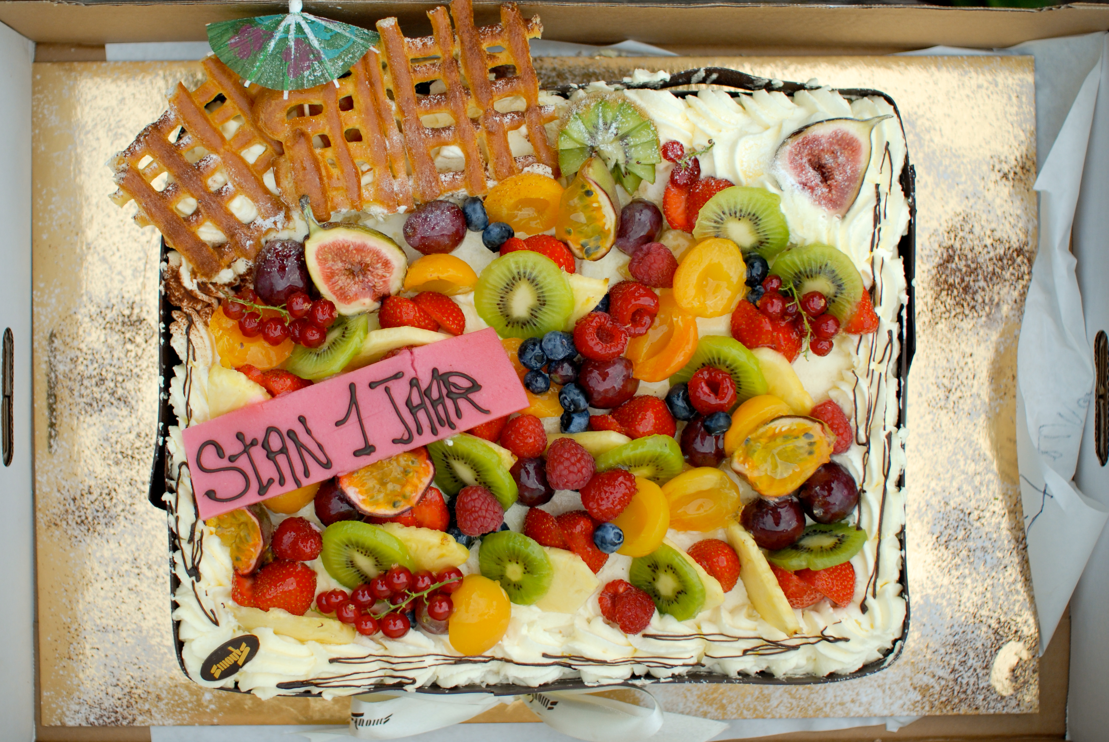 A rectangular birthday cake with whipped cream, fresh fruit, and a pink plaque reading "Stan 1 Jaar" in chocolate writing.