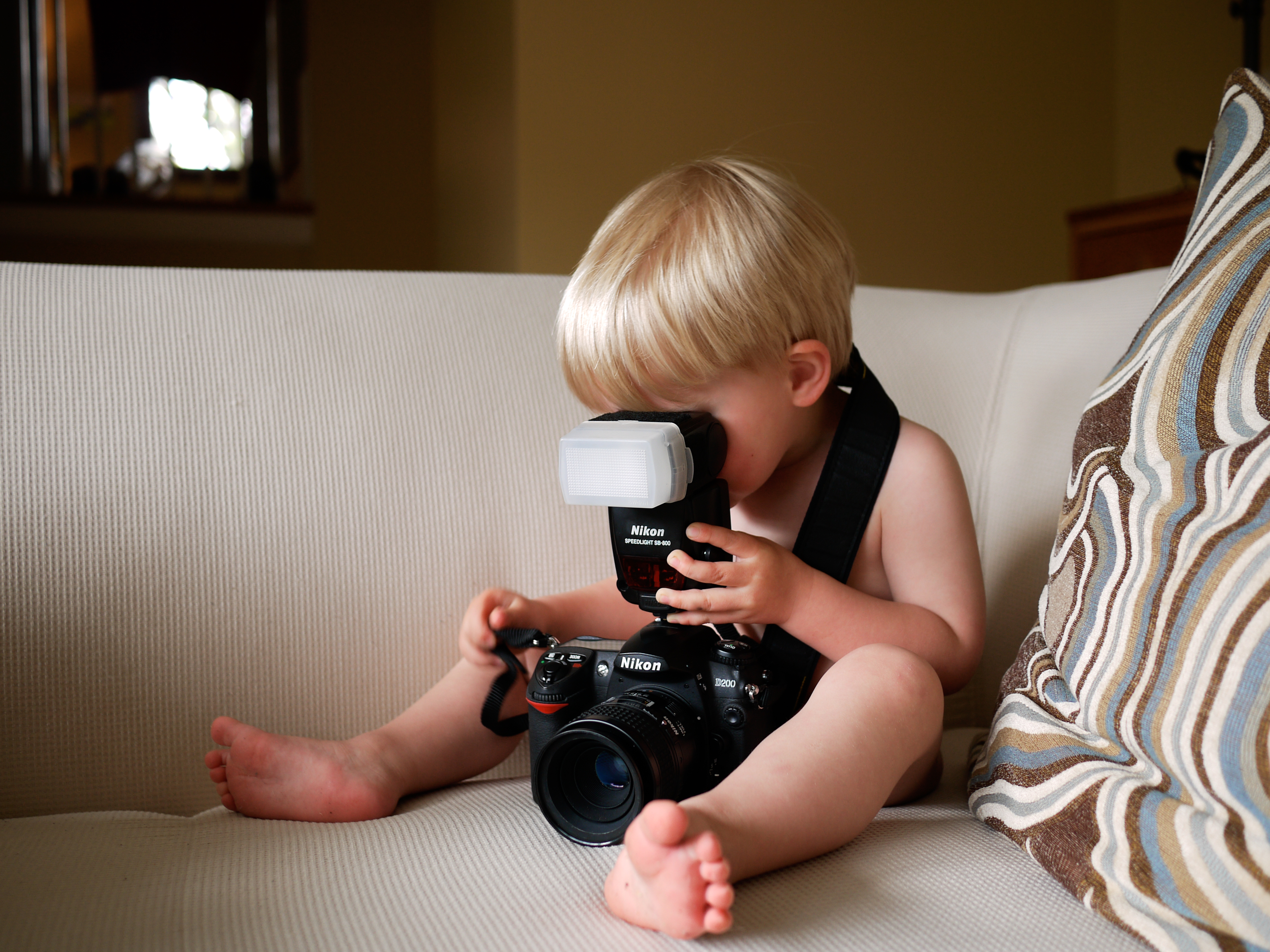 A young child sits on a couch, holding a large camera and looking through the viewfinder.