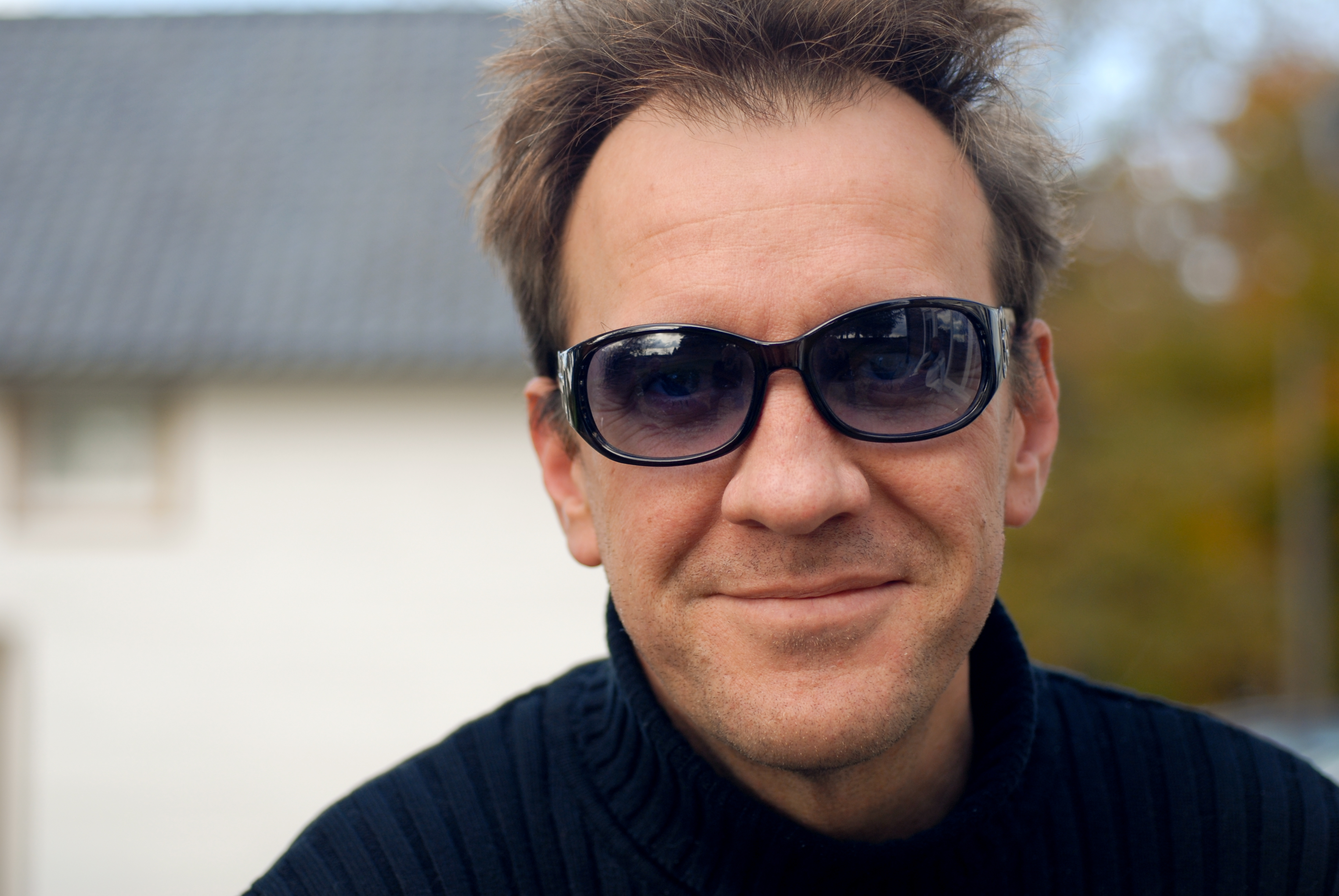 A man wearing dark sunglasses and a black sweater smiles slightly while looking at the camera outdoors.