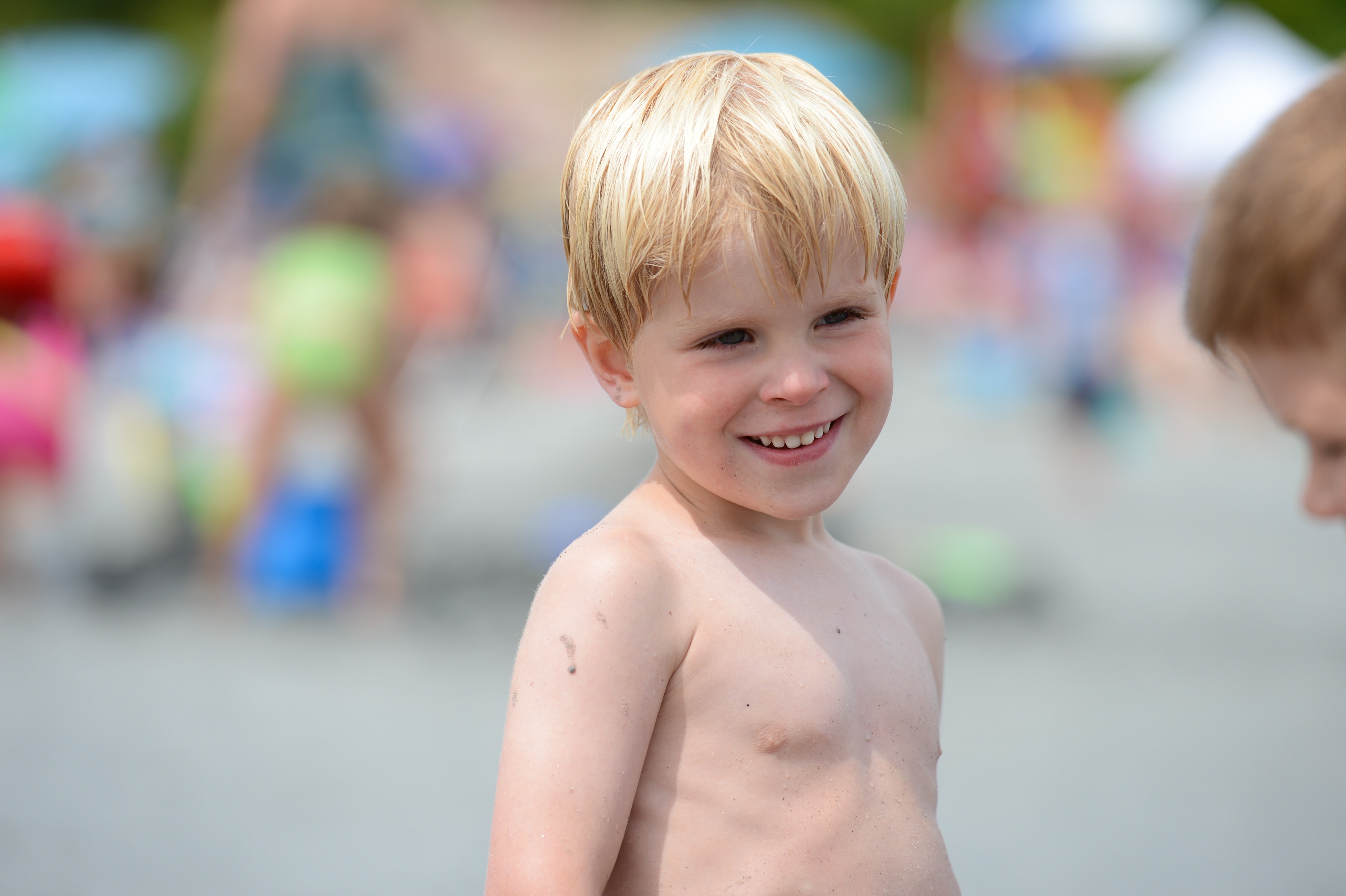 A young boy with wet blond hair smiles while standing shirtless on a busy beach.