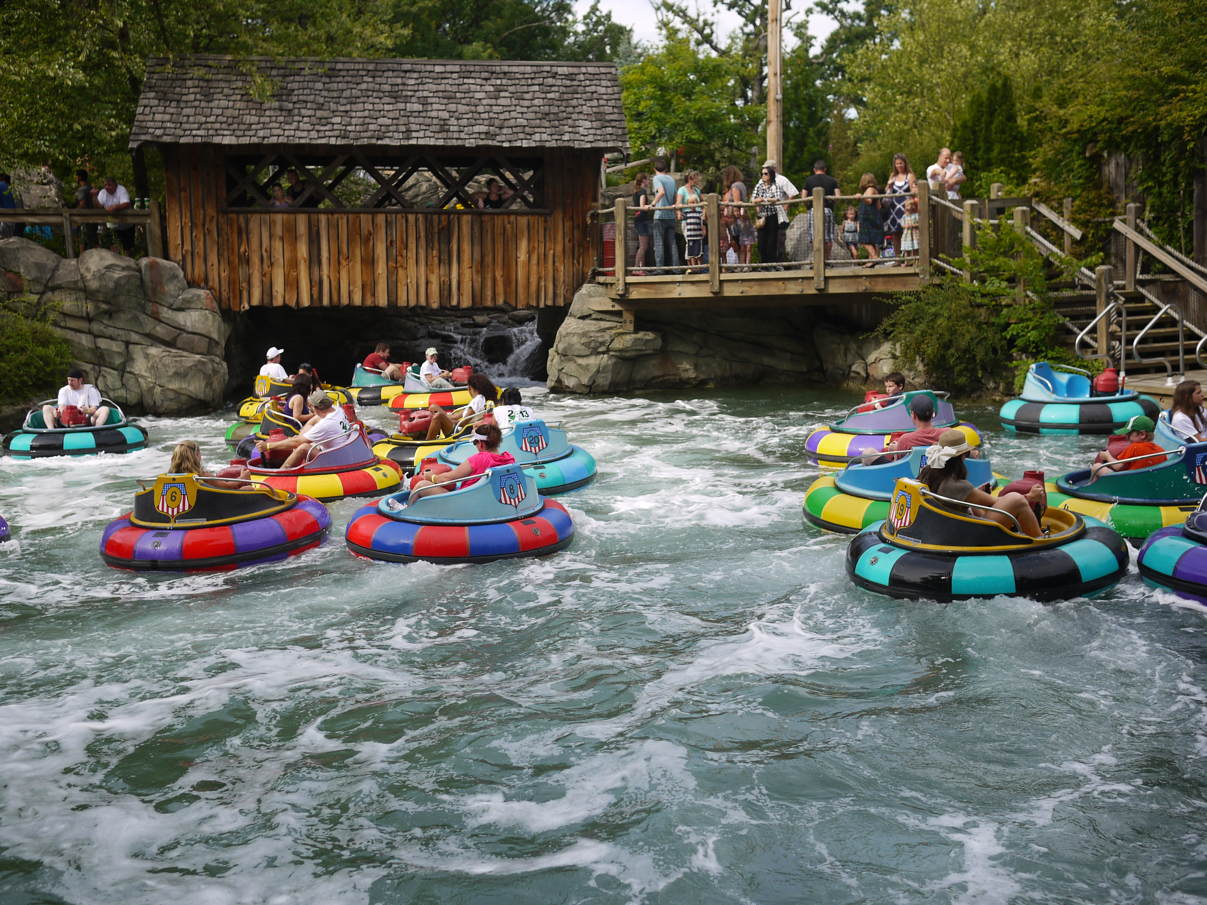 People in colorful bumper boats float on a water ride while others watch from a wooden bridge above.
