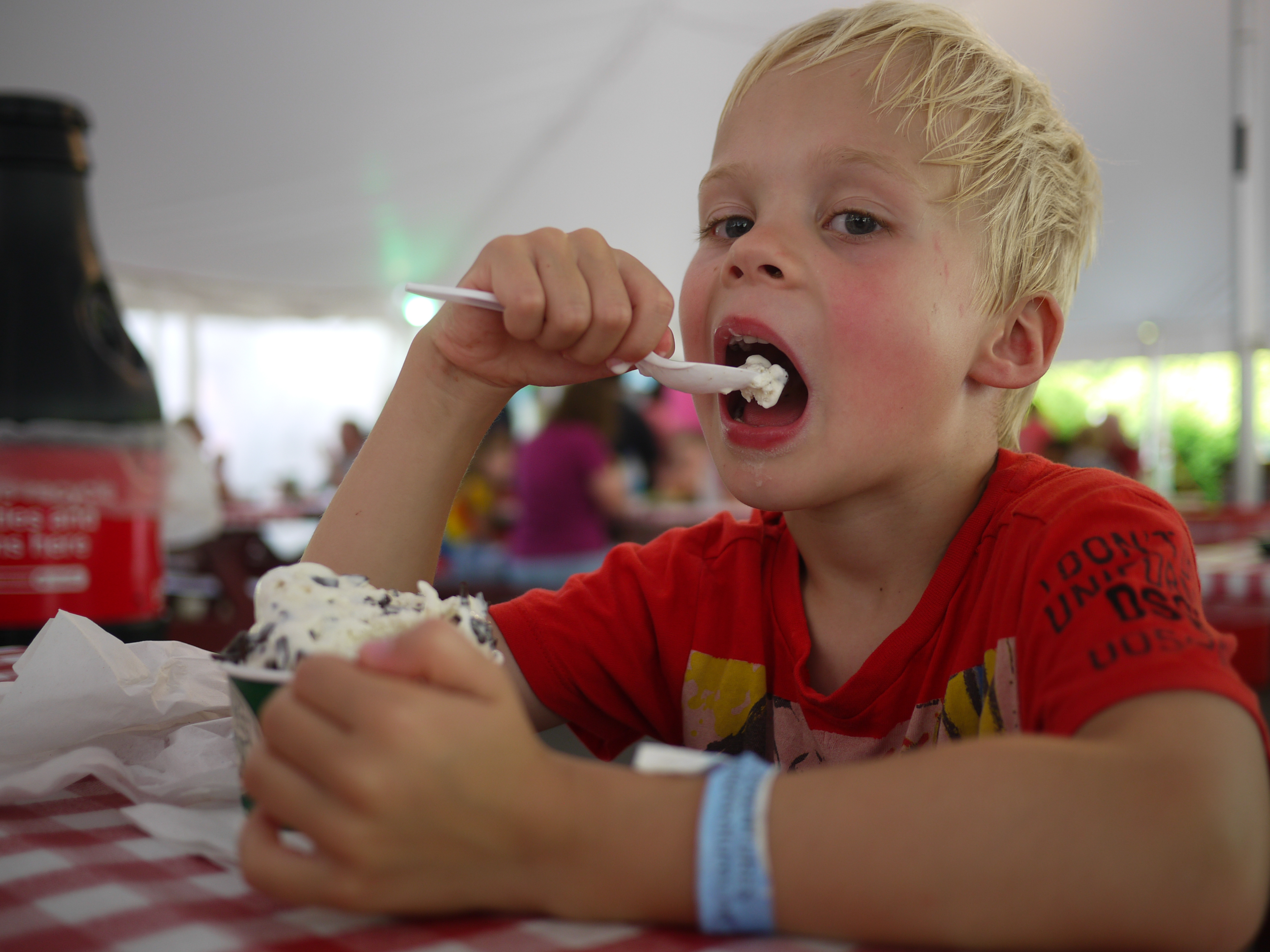 A young boy in a red shirt eats ice cream with a spoon at a table under a tent.