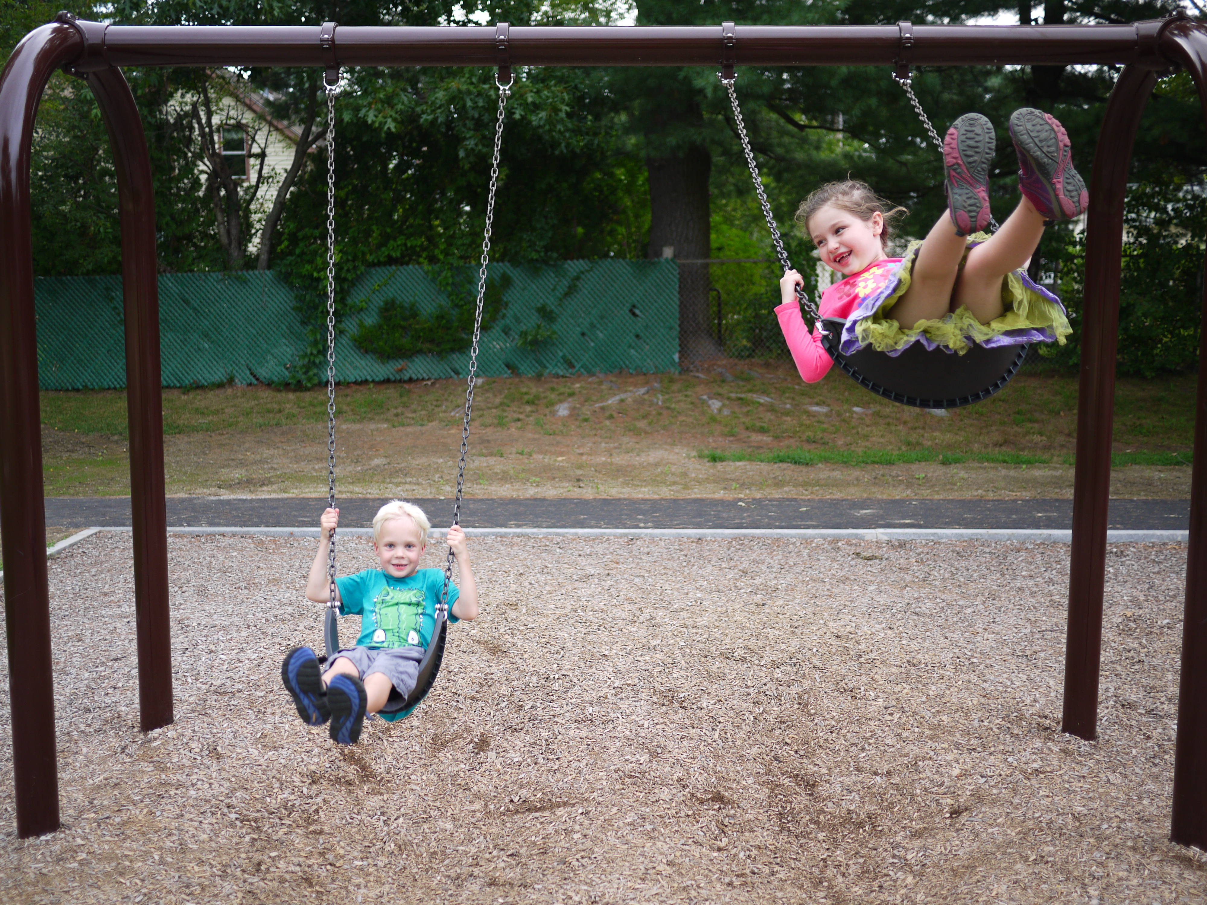 Two children swinging at a playground, one smiling and kicking their legs up while the other swings lower.