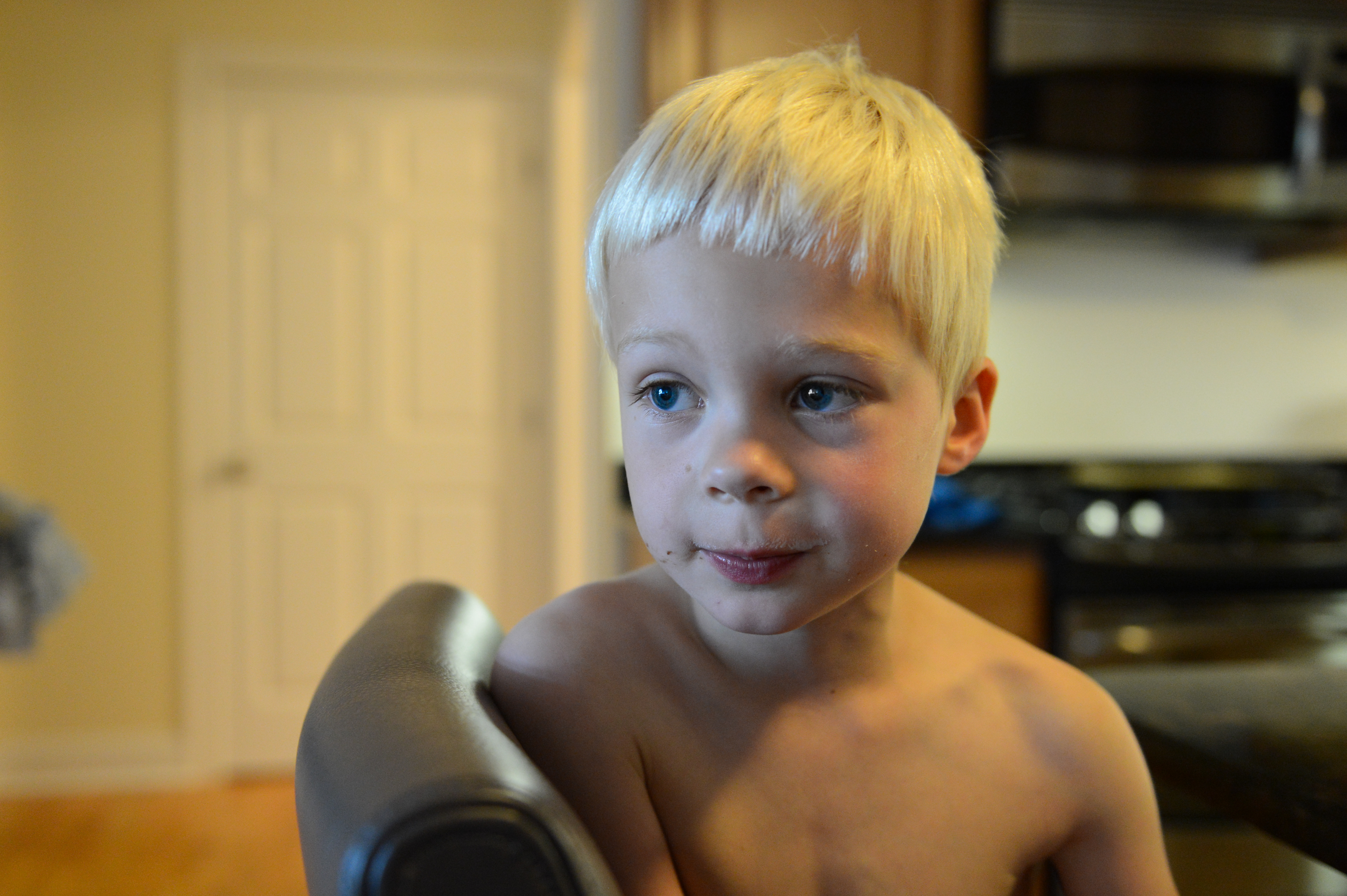 A young child with short blond hair sits shirtless on a chair, looking off to the side.