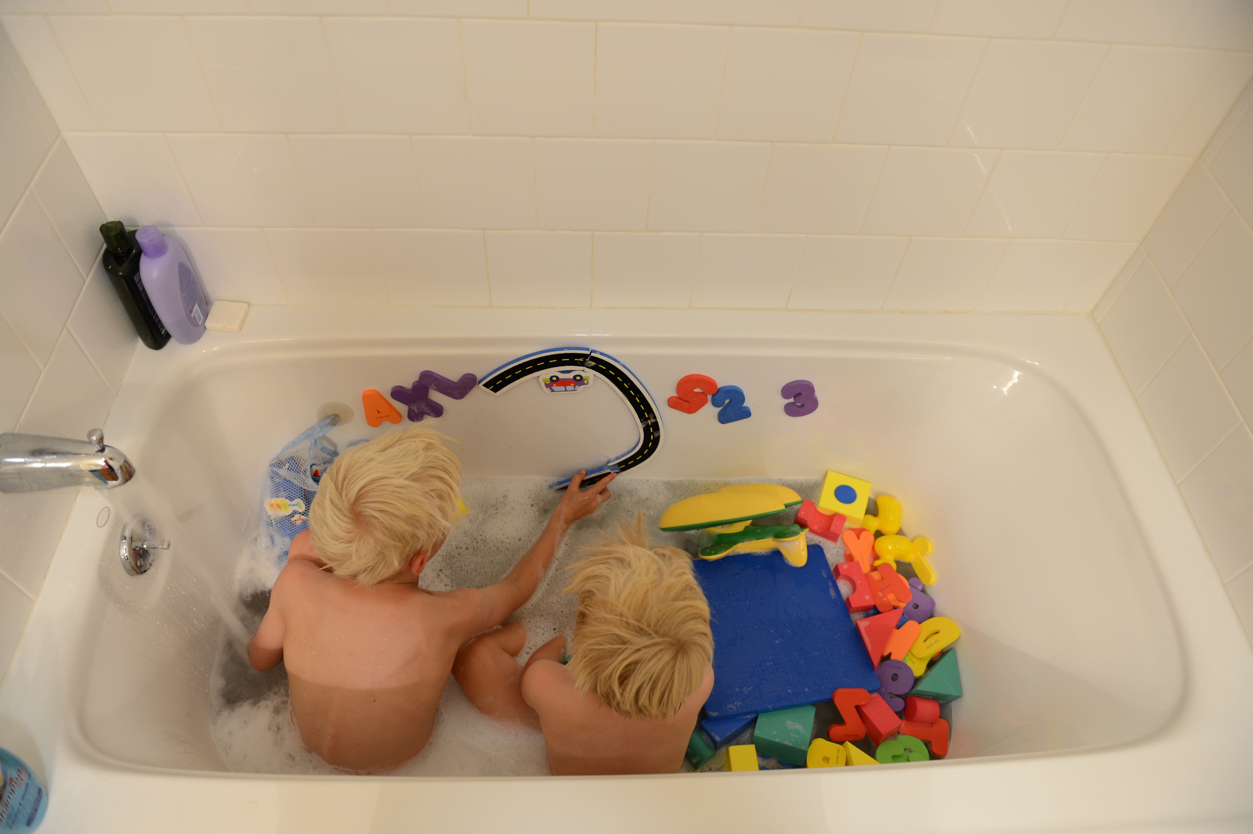 Two young children with blonde hair play with colorful foam letters and toys in a bathtub filled with water.