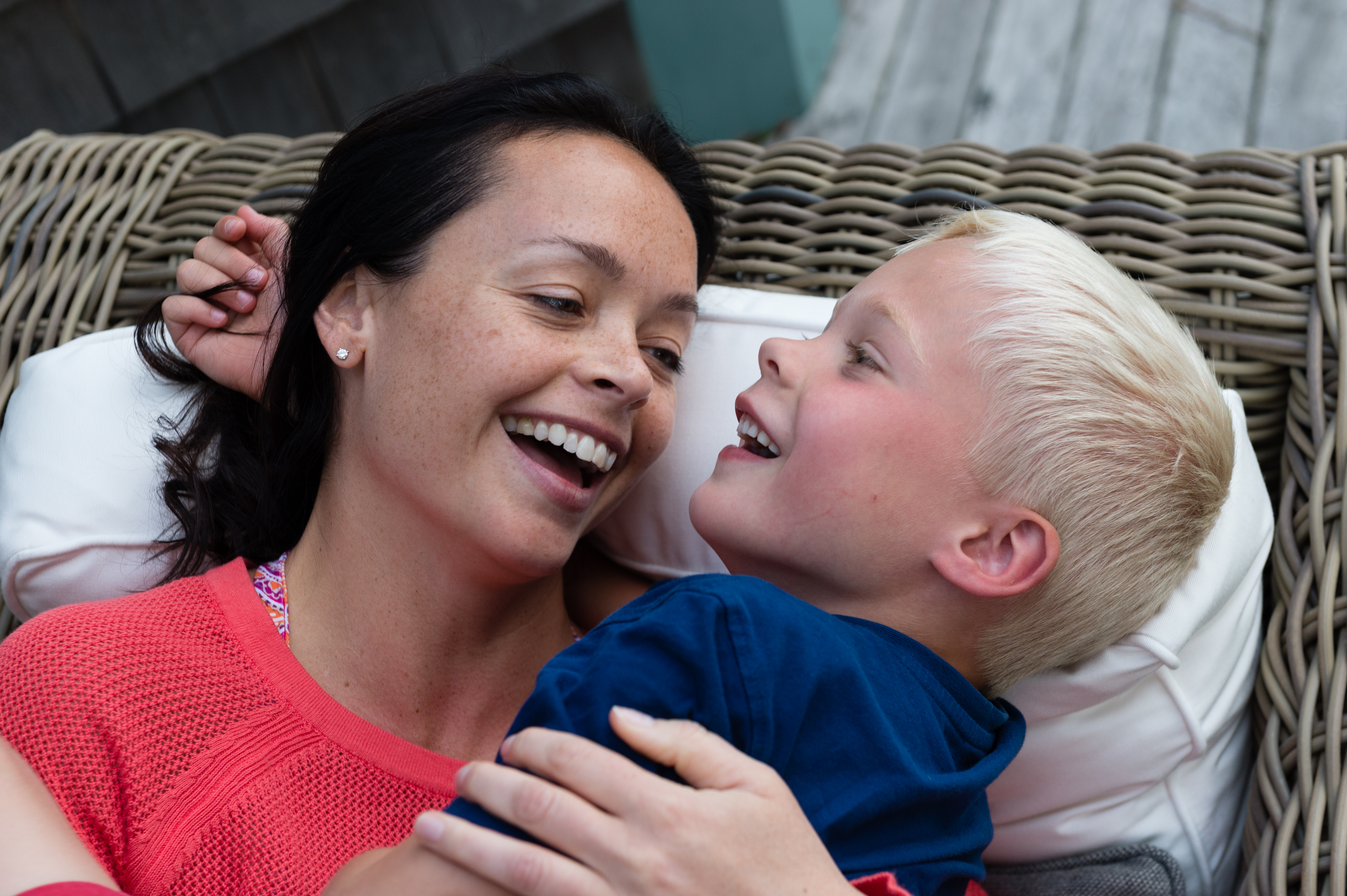 A woman and a young boy lie on a wicker lounge chair, smiling and laughing together.
