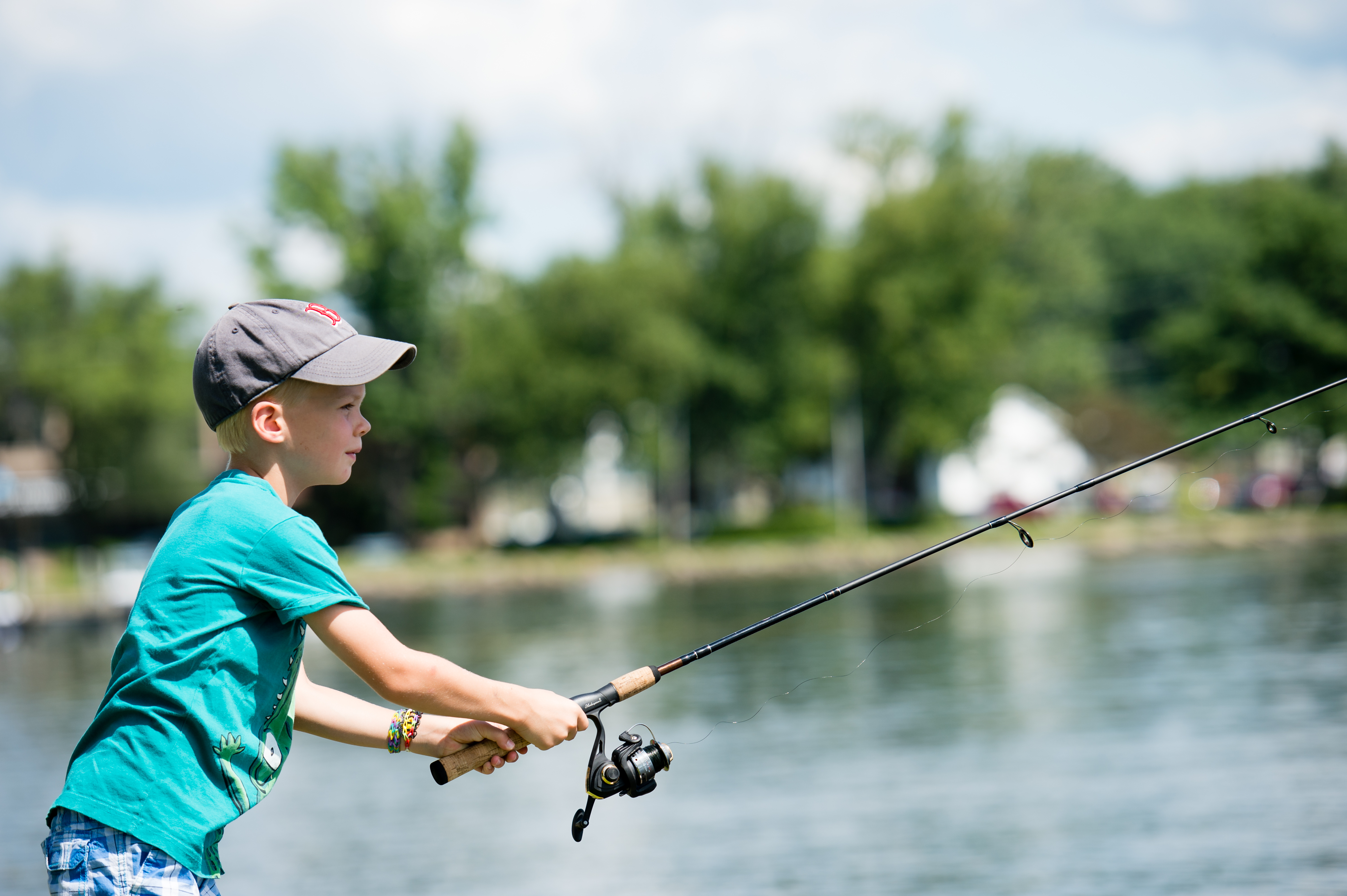 A young child wearing a cap and t-shirt holds a fishing rod while casting a line into the water.