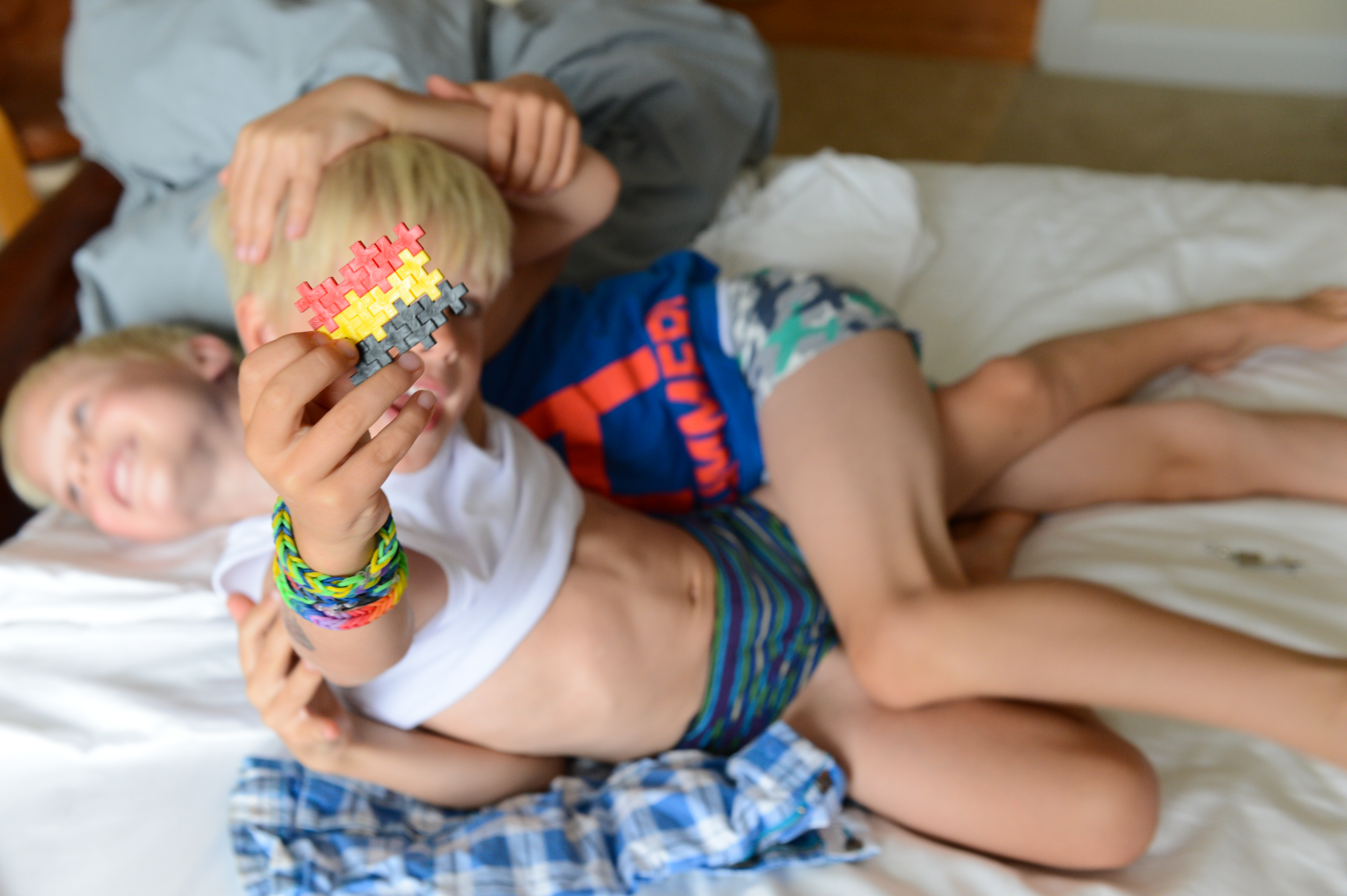 Two children playfully wrestle on a bed while one holds up a small colorful puzzle piece.