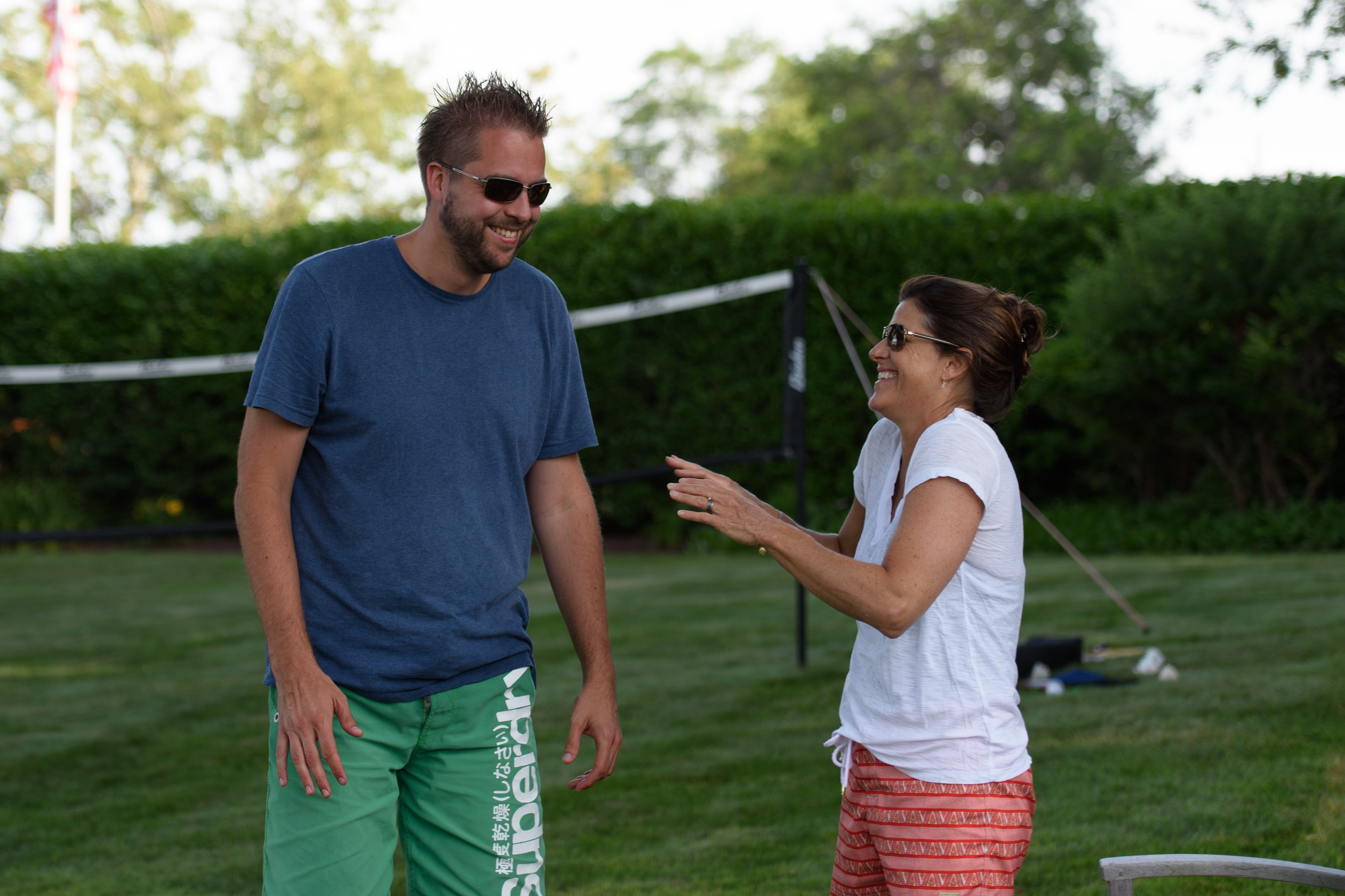 A man and a woman laugh together outside near a volleyball net on a grassy lawn.