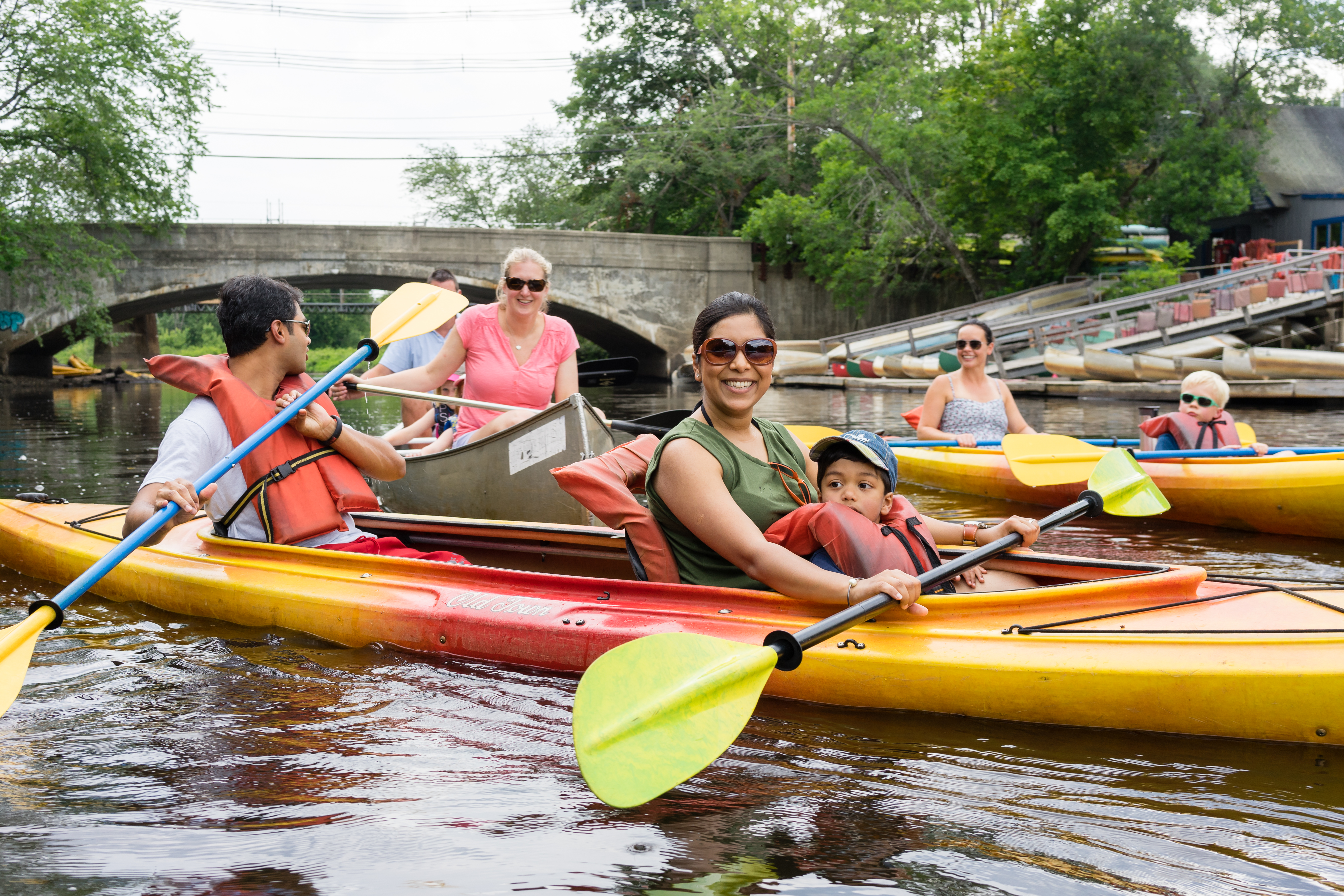 A group of people kayaking on a calm river, with a woman and child smiling in the foreground.