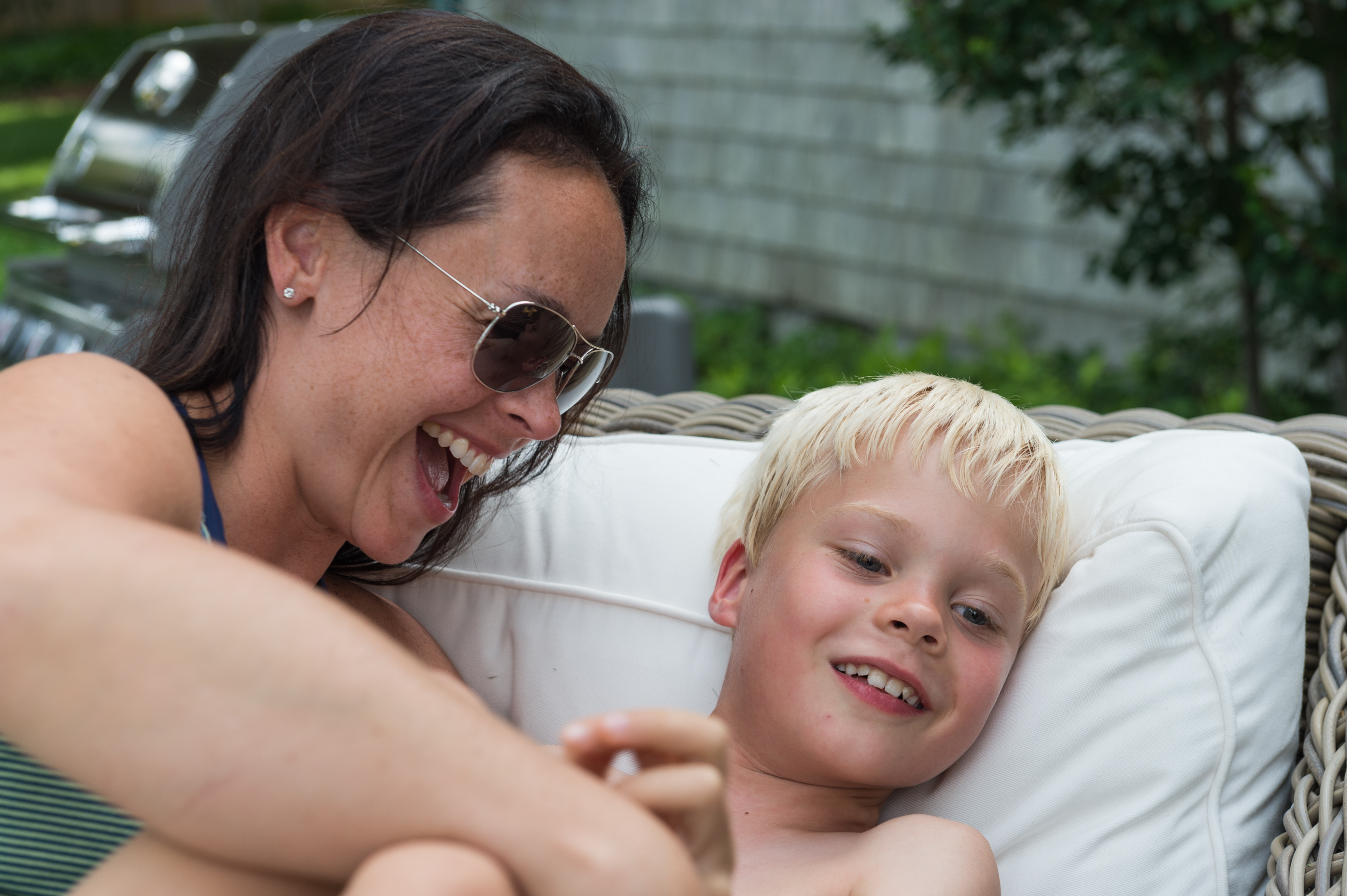 A woman laughs while playfully tickling a smiling young boy as he relaxes on a cushioned outdoor chair.