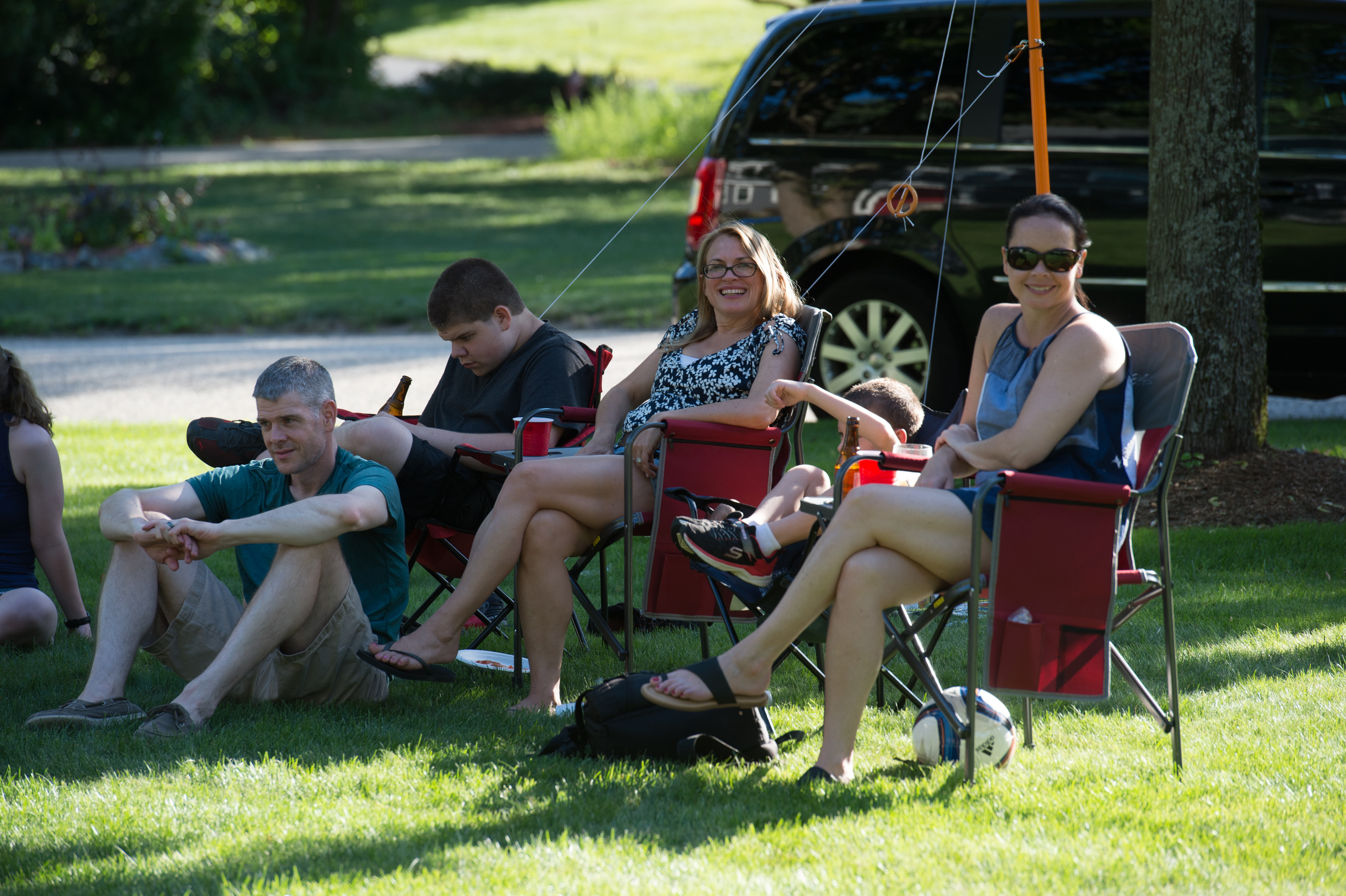 A group of people sitting in folding chairs on the grass, relaxing in a backyard on a sunny day.