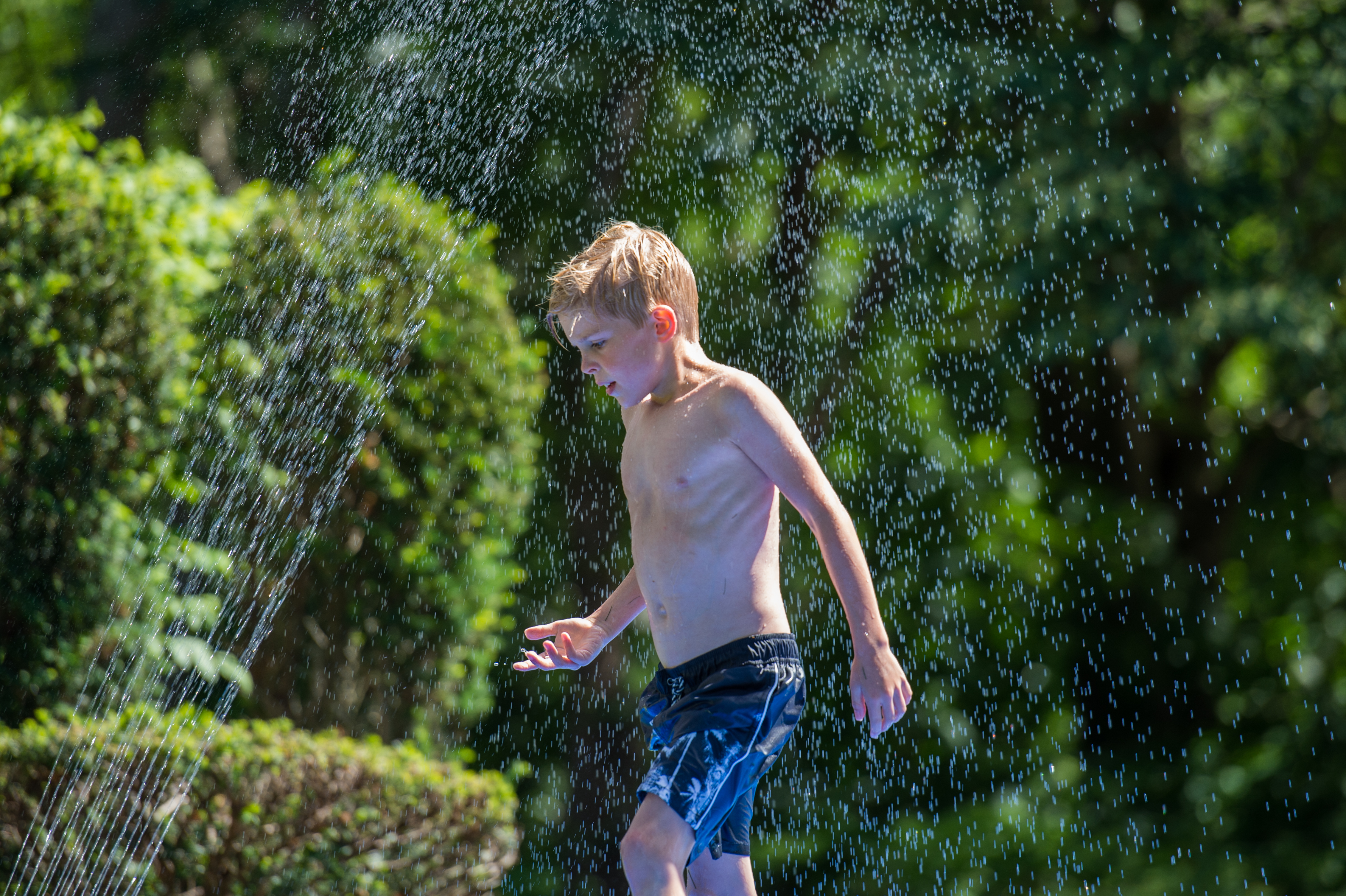 A young boy in swim trunks walks through a water spray, enjoying a summer day outdoors.