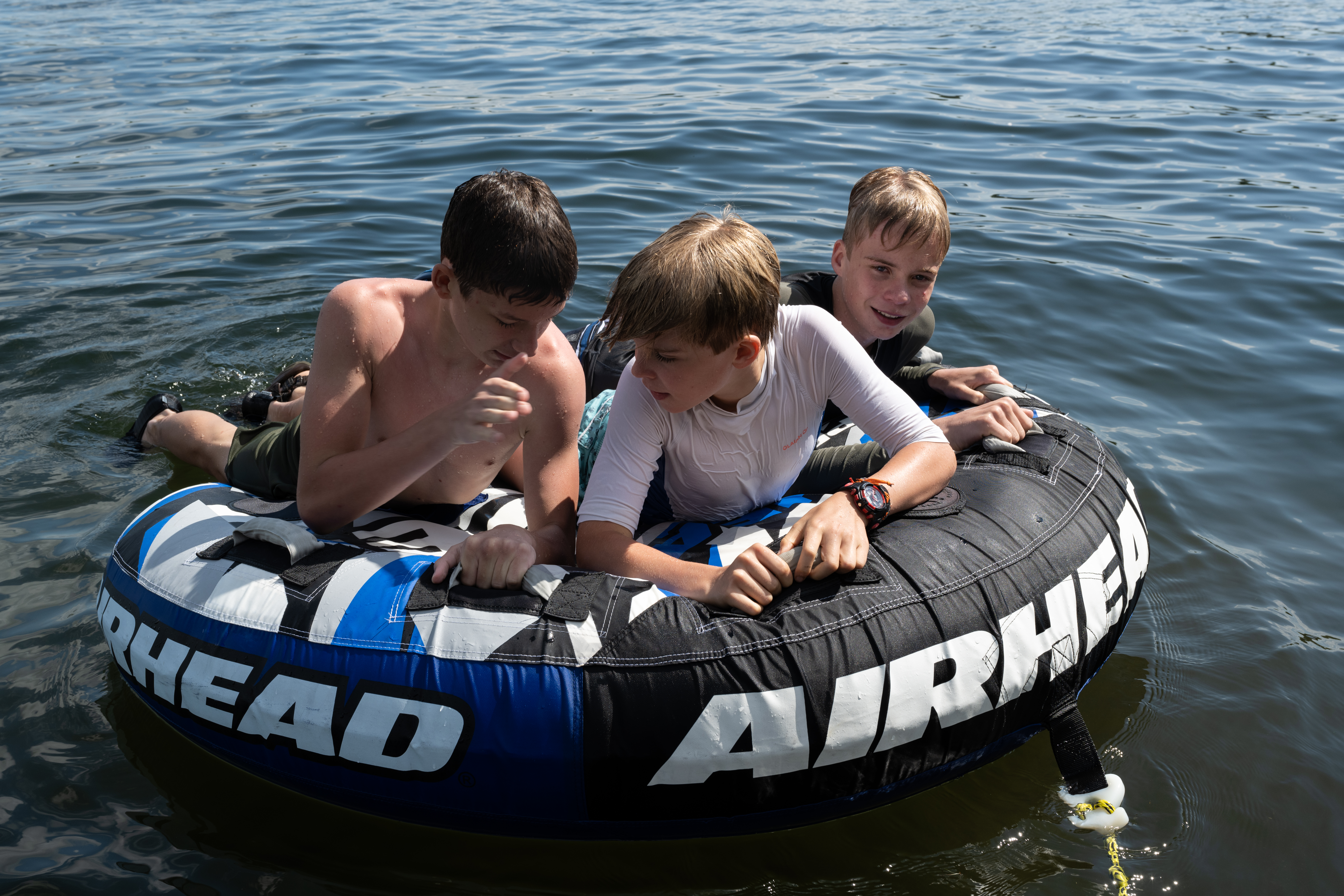 Three boys lay on a large inflatable tube floating on a lake, holding on and chatting.