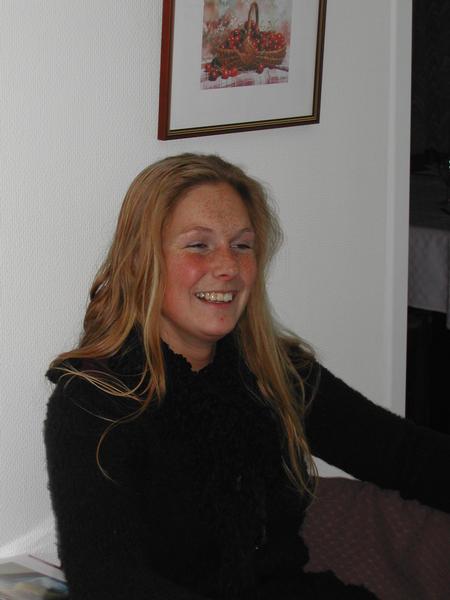 A smiling woman with long hair sits indoors, wearing a black sweater.