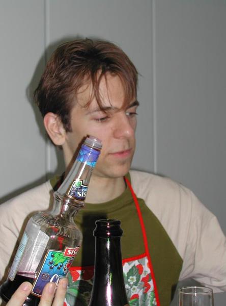 A man wearing an apron looks at a bottle while holding it during a dinner gathering.
