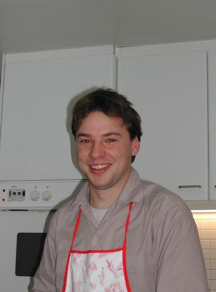 A smiling person wearing an apron stands in a kitchen, looking at the camera.