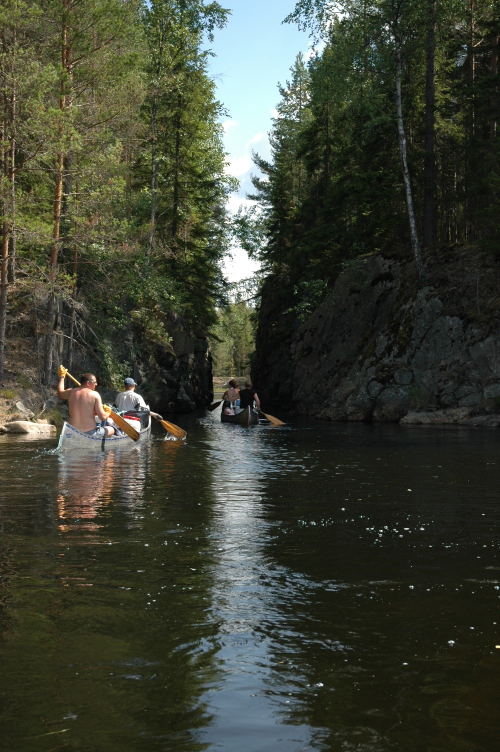 People paddling canoes through a narrow waterway surrounded by tall cliffs and dense green trees.