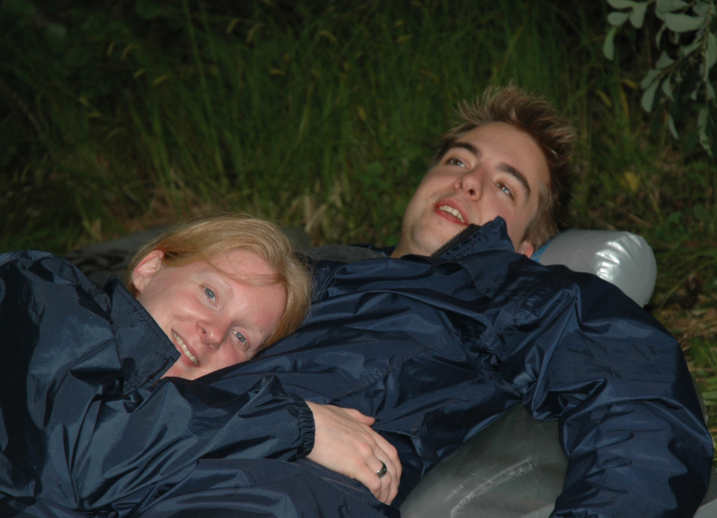 Two people in matching jackets lying down outdoors, one smiling at the camera while resting on the other's chest.