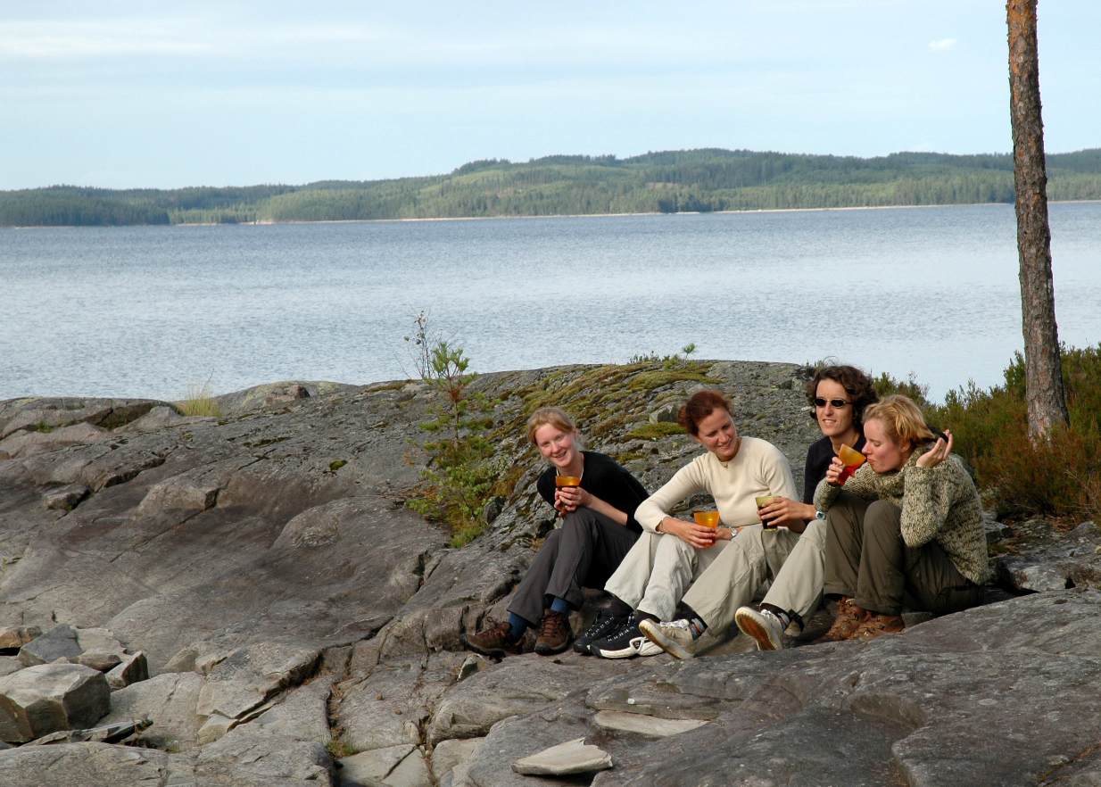 Four women sit on rocky ground near a lake, smiling and drinking from glasses.