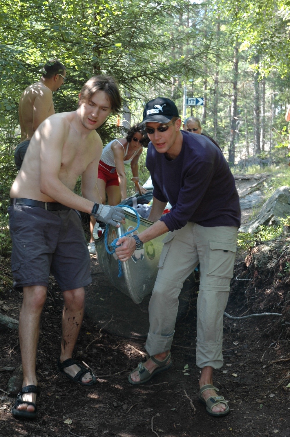 Two people lift a canoe over rough ground while others assist in a wooded area.