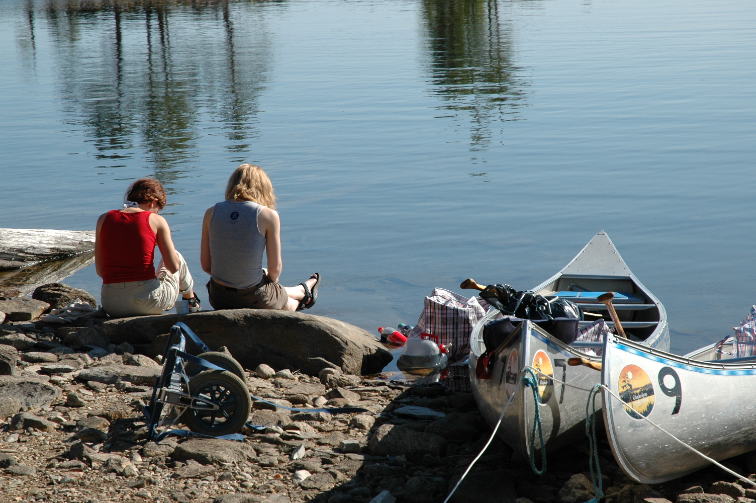 Two people sit on a rock by the water, reading a book together.