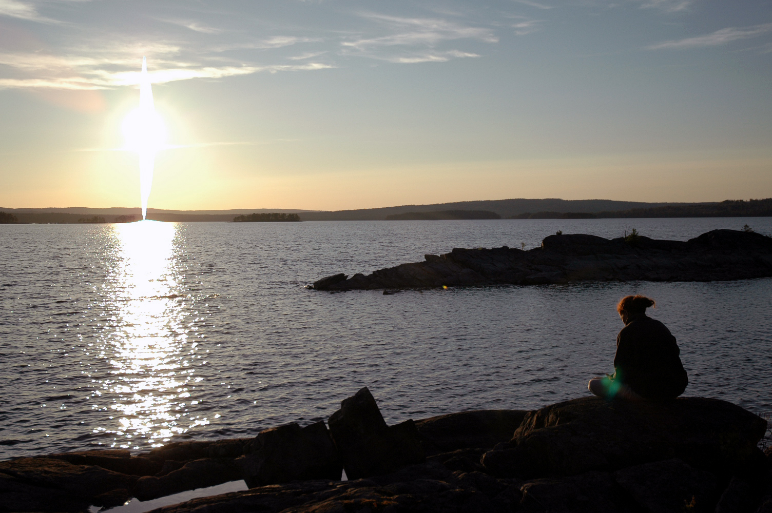 A person sits on a rocky shore, looking at the water during sunset.