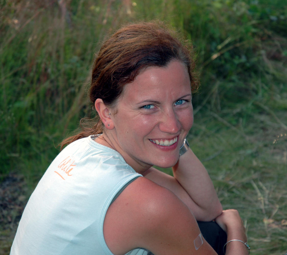 A woman with brown hair and blue eyes smiles while sitting outdoors on a grassy area.