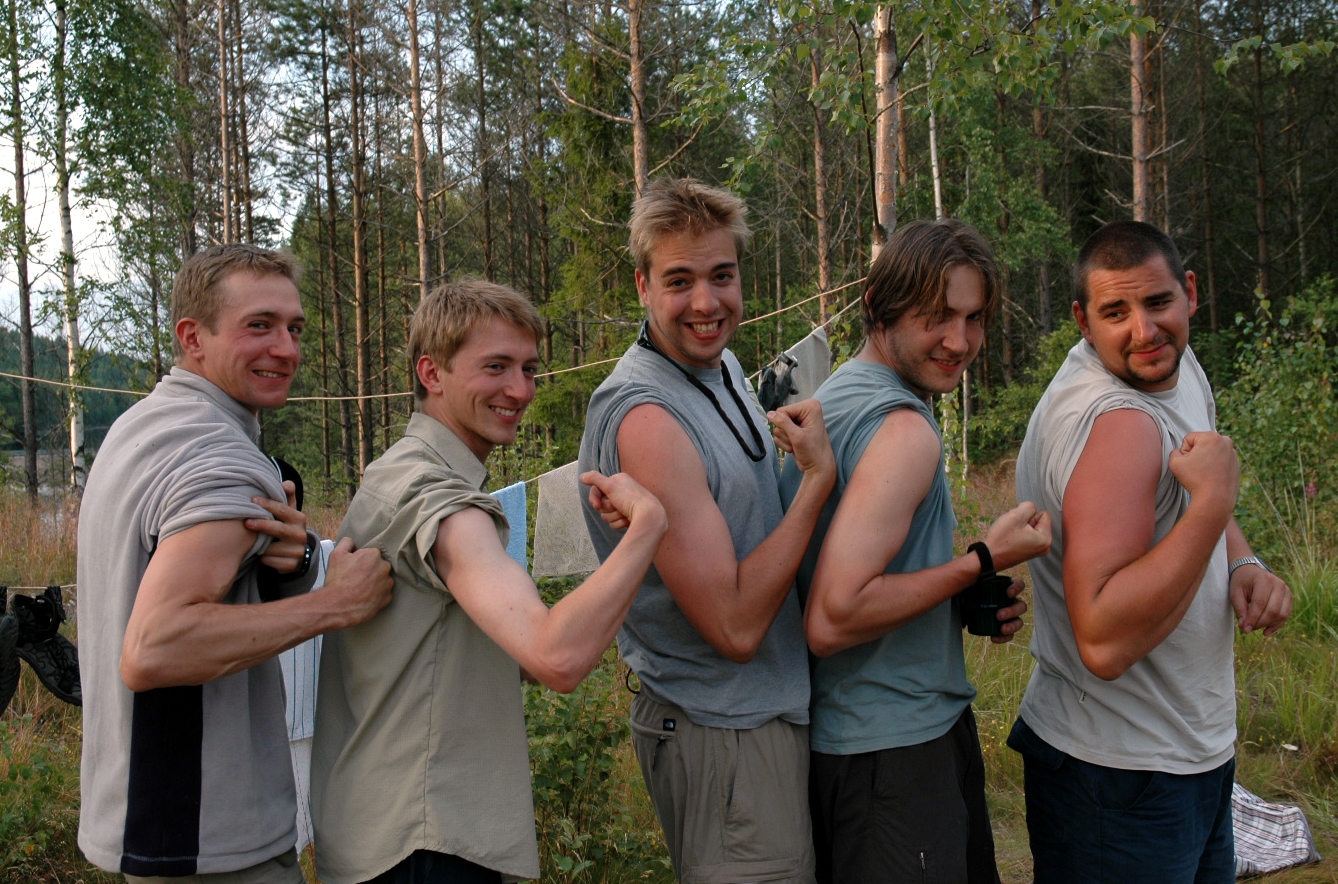 Five young men stand in a row outdoors, smiling and flexing their arms while pulling up their sleeves.
