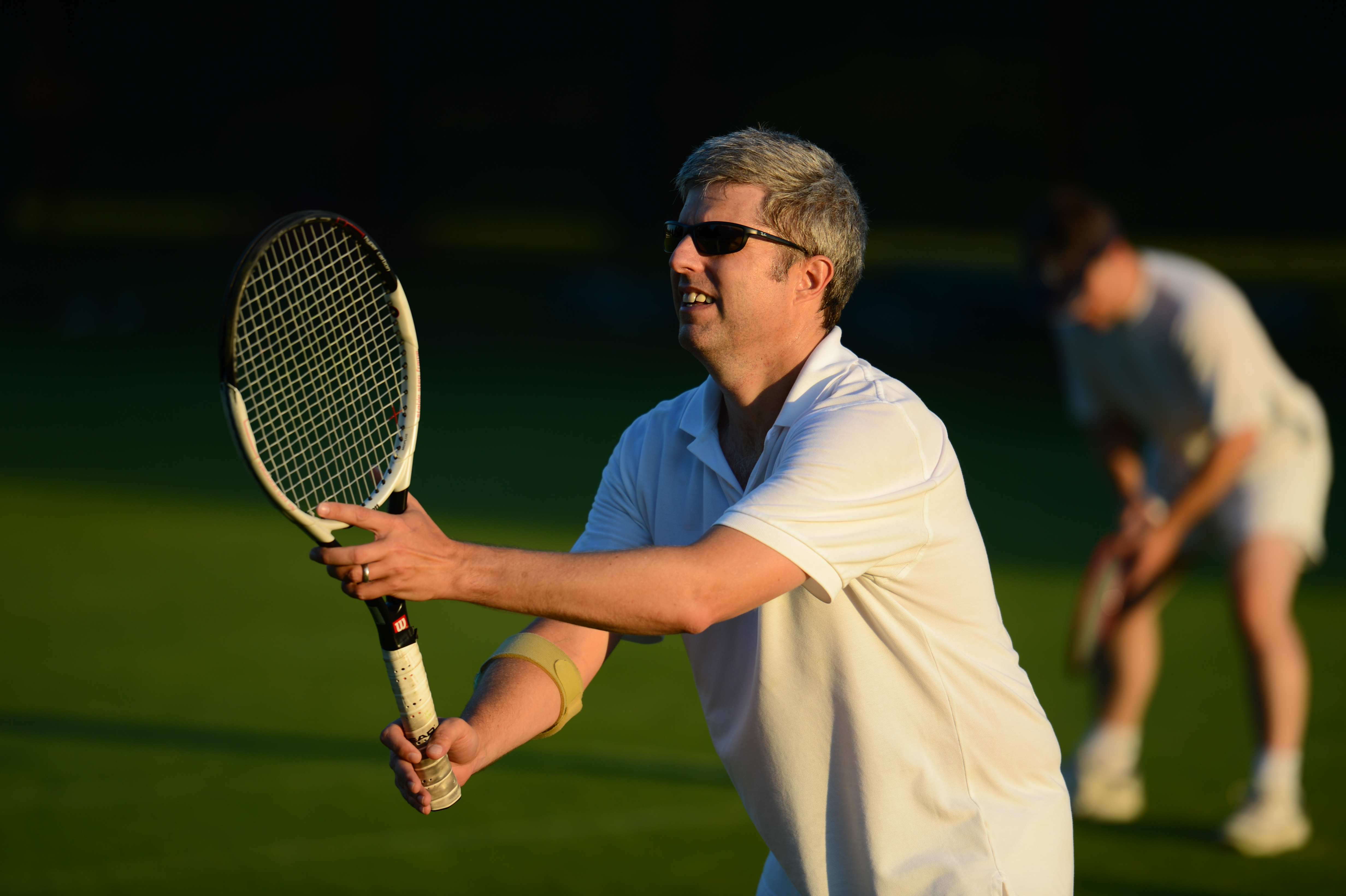 A man in a white outfit and sunglasses prepares to hit a tennis ball with his racket.