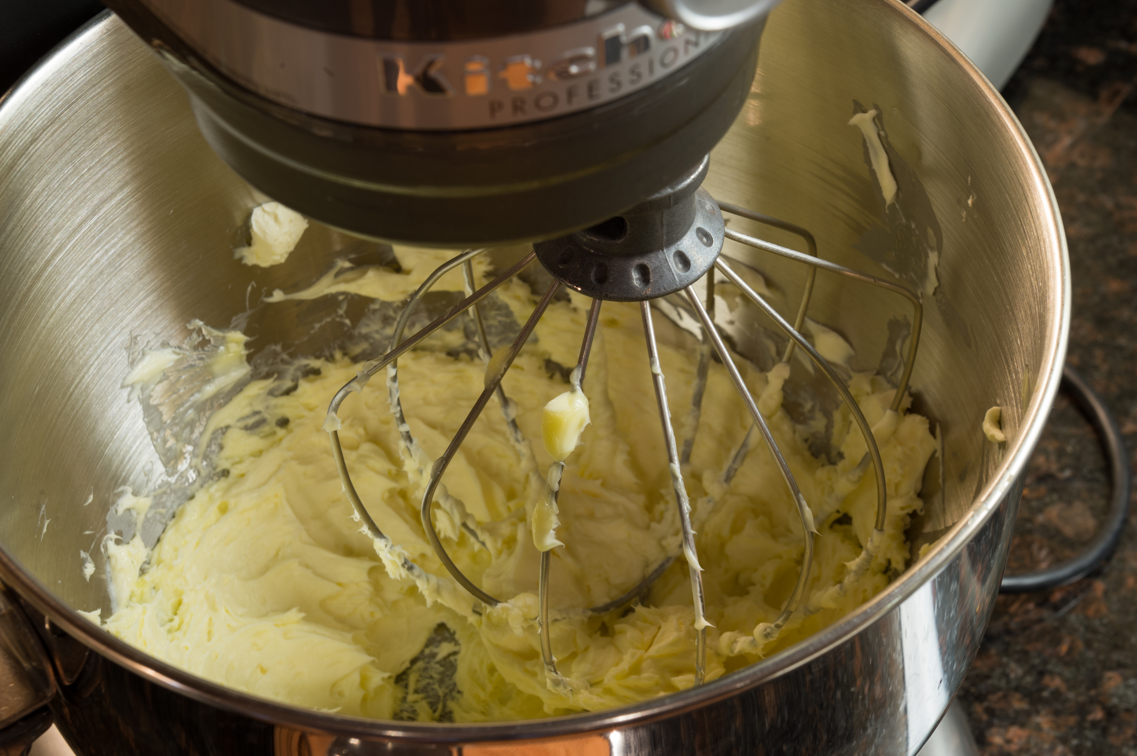 A stand mixer with a whisk attachment is mixing butter in a stainless steel bowl.