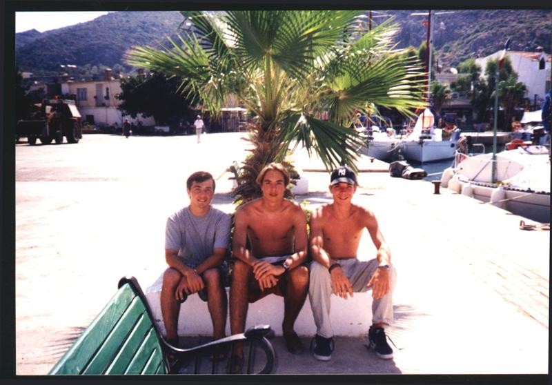 Three young men sit on a low wall near a palm tree, smiling at the camera in a sunny marina.