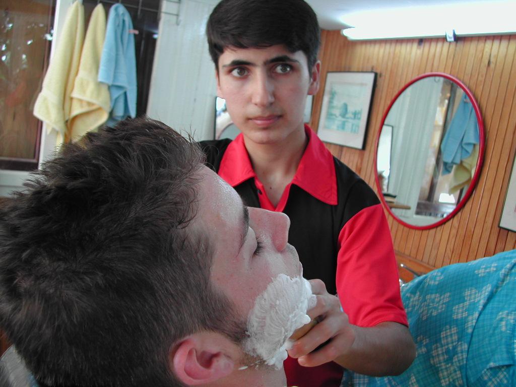 A barber in a red and black shirt applies shaving cream to a customer's face in a barbershop.