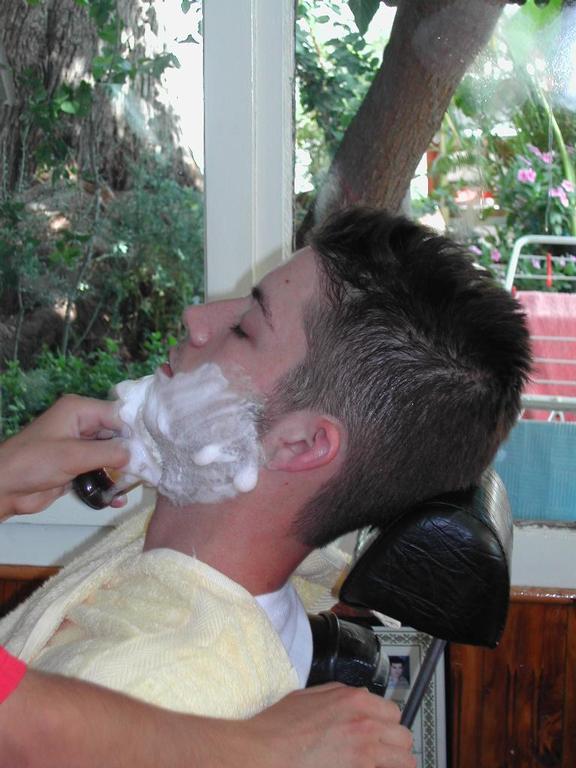 A barber shaves a young man's face with shaving cream while he reclines in a chair.