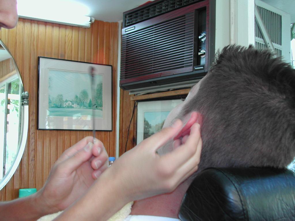 A barber cleans a customer's ear with a tool while the customer sits in a chair.