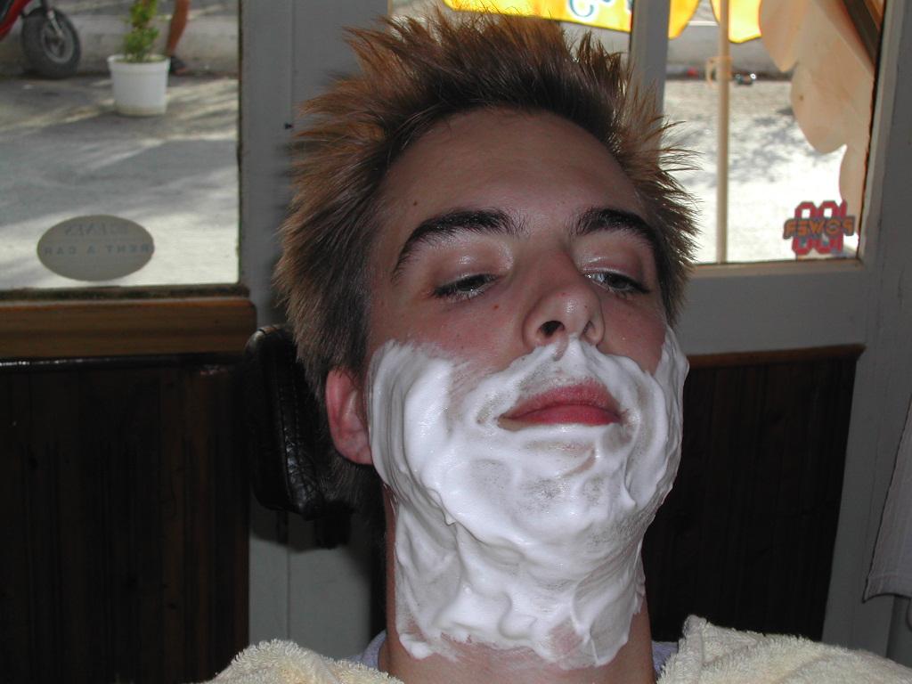 A person sits in a barber chair with shaving cream on their face, preparing for a shave.